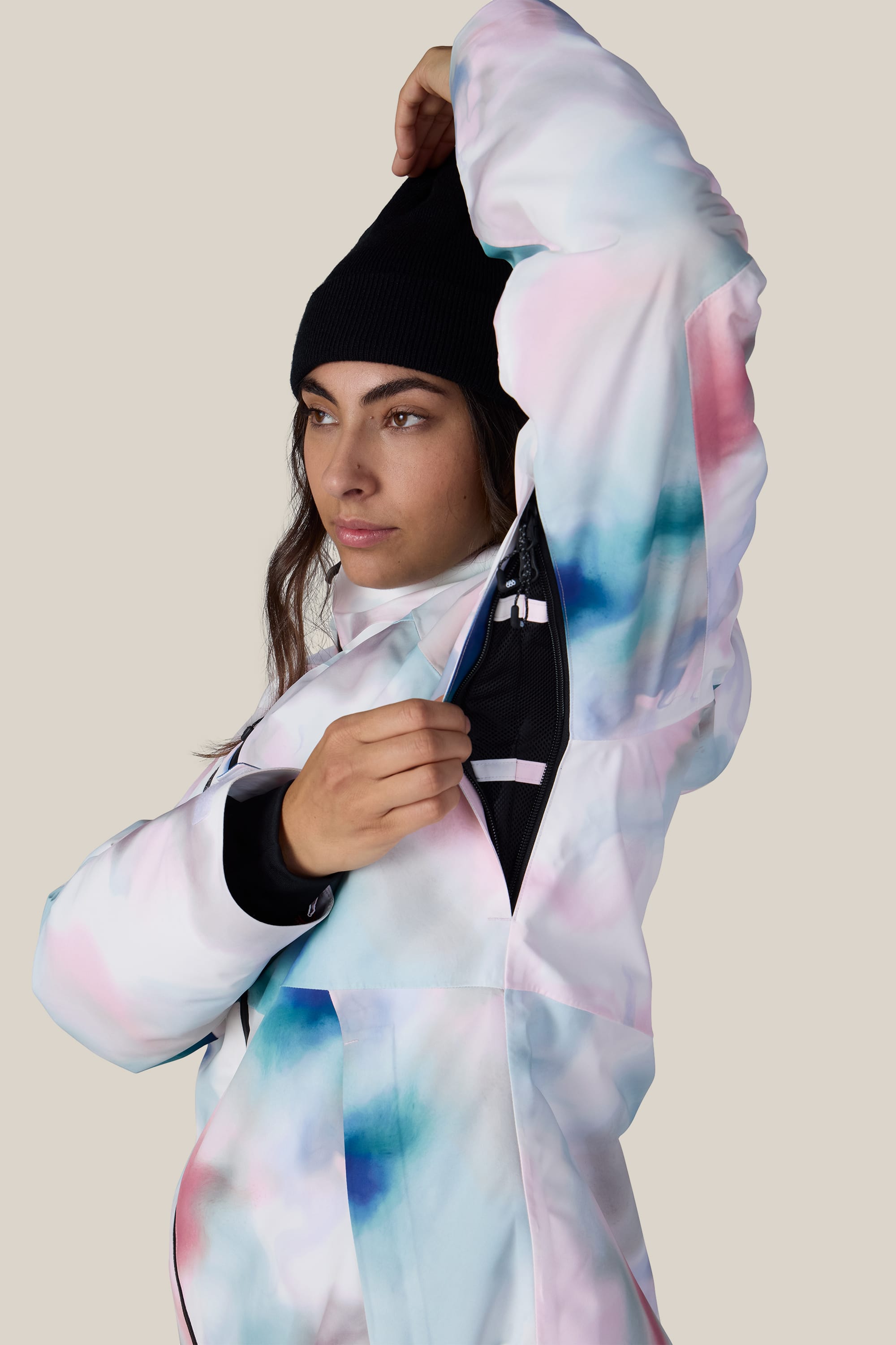 Wearing the 686 Women’s Hydra™ Thermagraph® Jacket, a person in a black beanie lifts one arm and unzips the underarm ventilation opening, revealing the black inner lining, against a plain beige background.