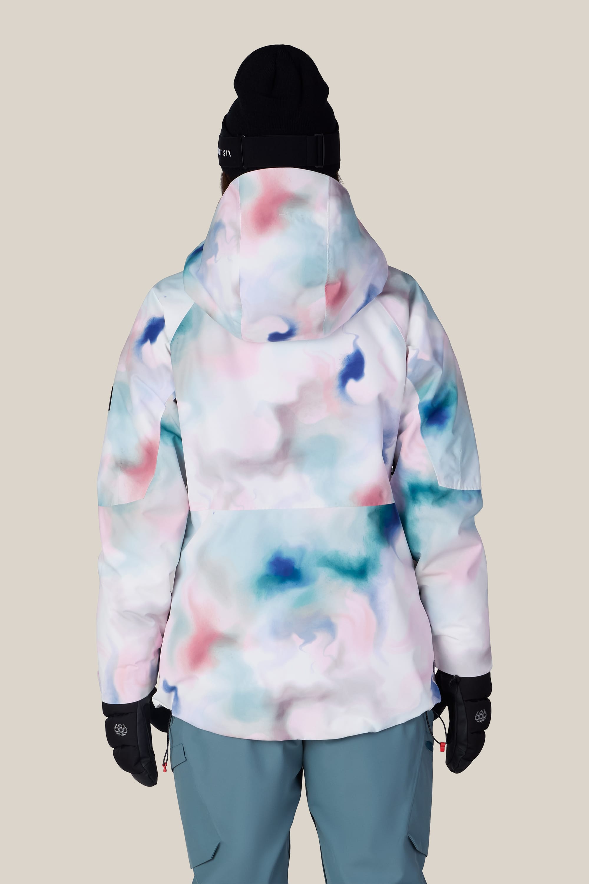A person wearing the 686 Women’s Hydra™ Thermagraph® Jacket by 686, featuring a pastel watercolor pattern, along with blue pants, black gloves, and a black beanie, stands with their back to the camera against a plain beige background.