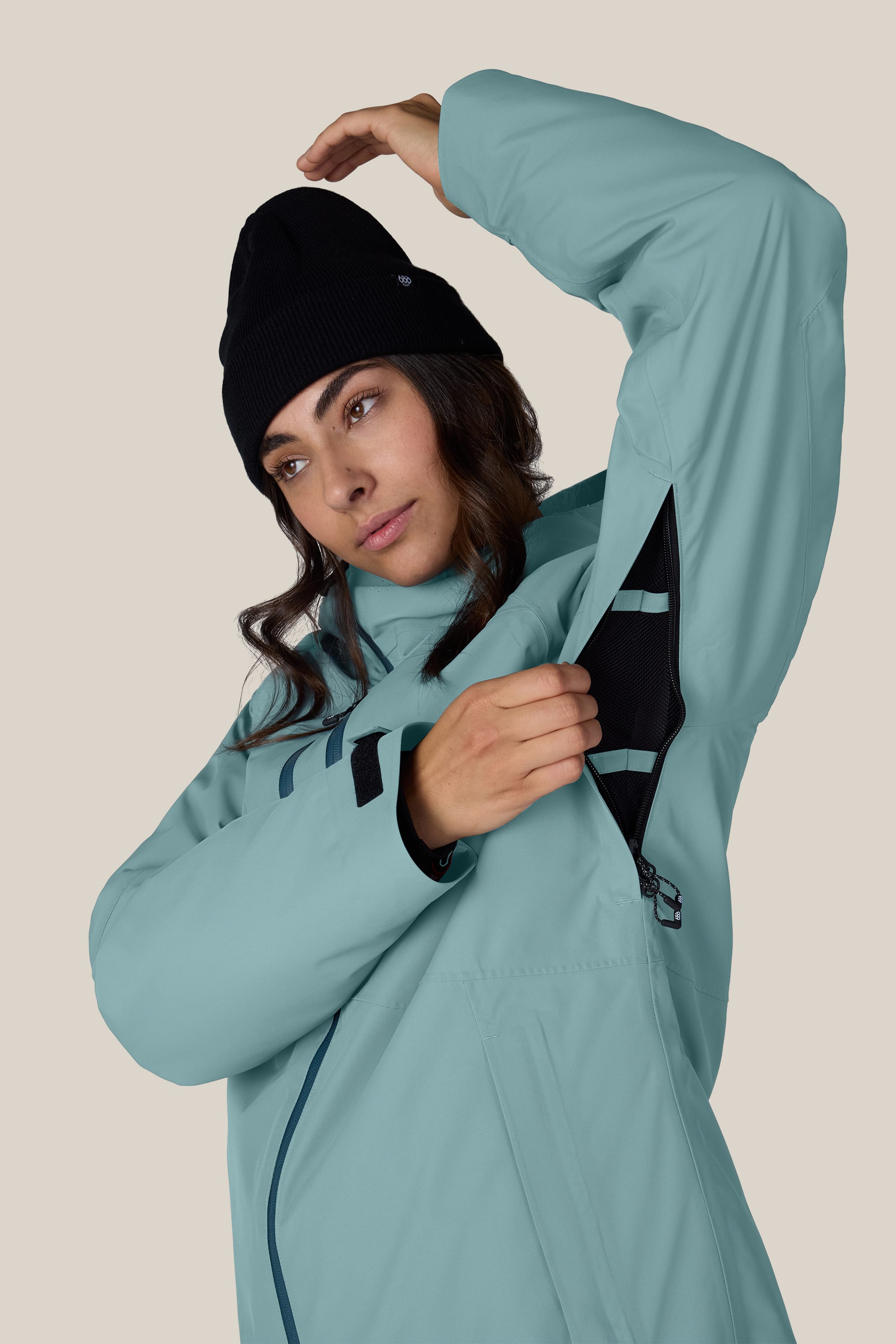 Wearing the 686 Women’s Hydra™ Thermagraph® Jacket in light blue, a person with a black beanie unzips an underarm ventilation opening, showcasing the jacket’s features.