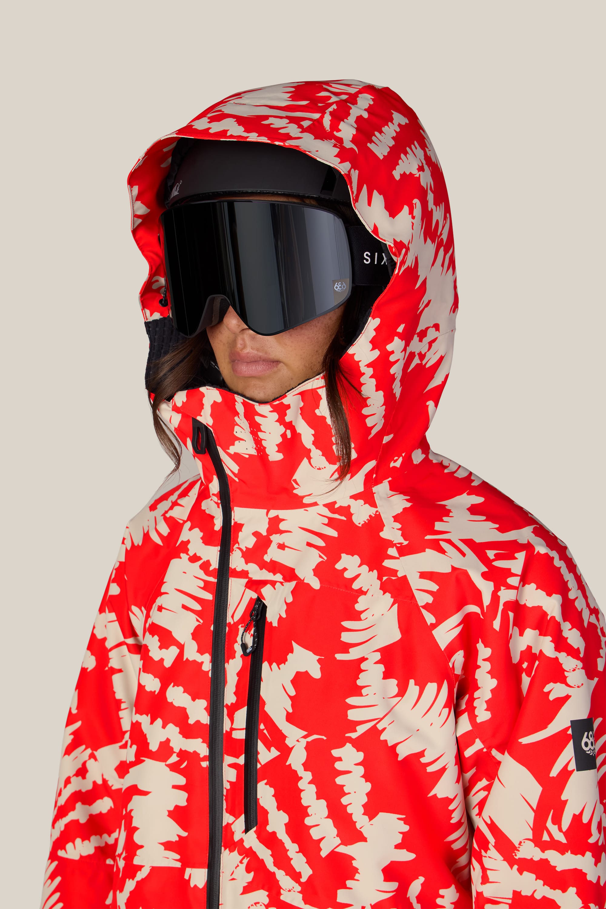 A person wearing the 686 Women’s Hydra™ Thermagraph® Jacket in a red and cream pattern, paired with large black ski goggles, faces forward against a neutral background.