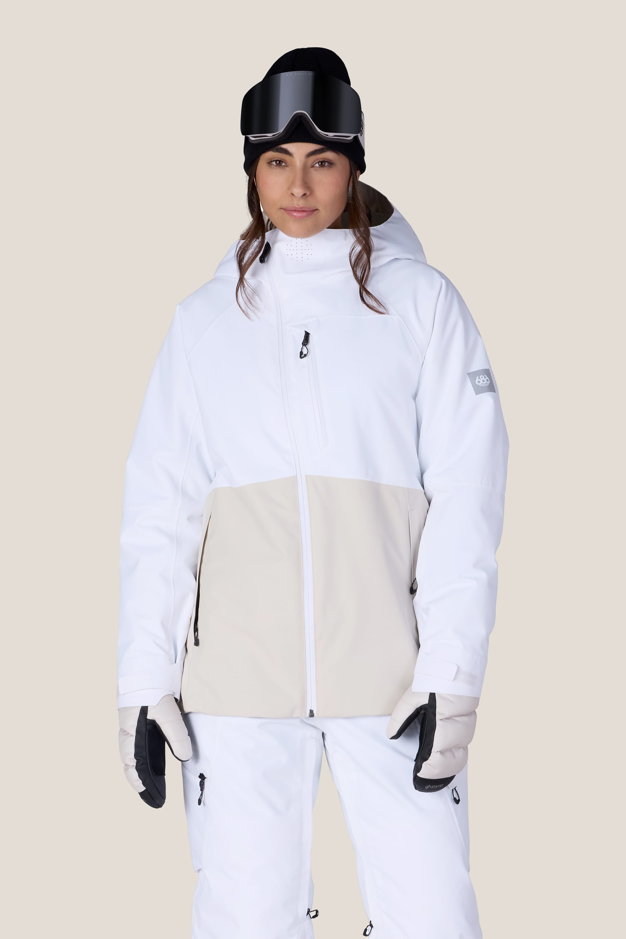 A person wearing the 686 Women’s Hydra™ Thermagraph® Jacket by 686 in white and beige, paired with white snow pants, gloves, a black beanie, and ski goggles stands facing forward against a plain background.