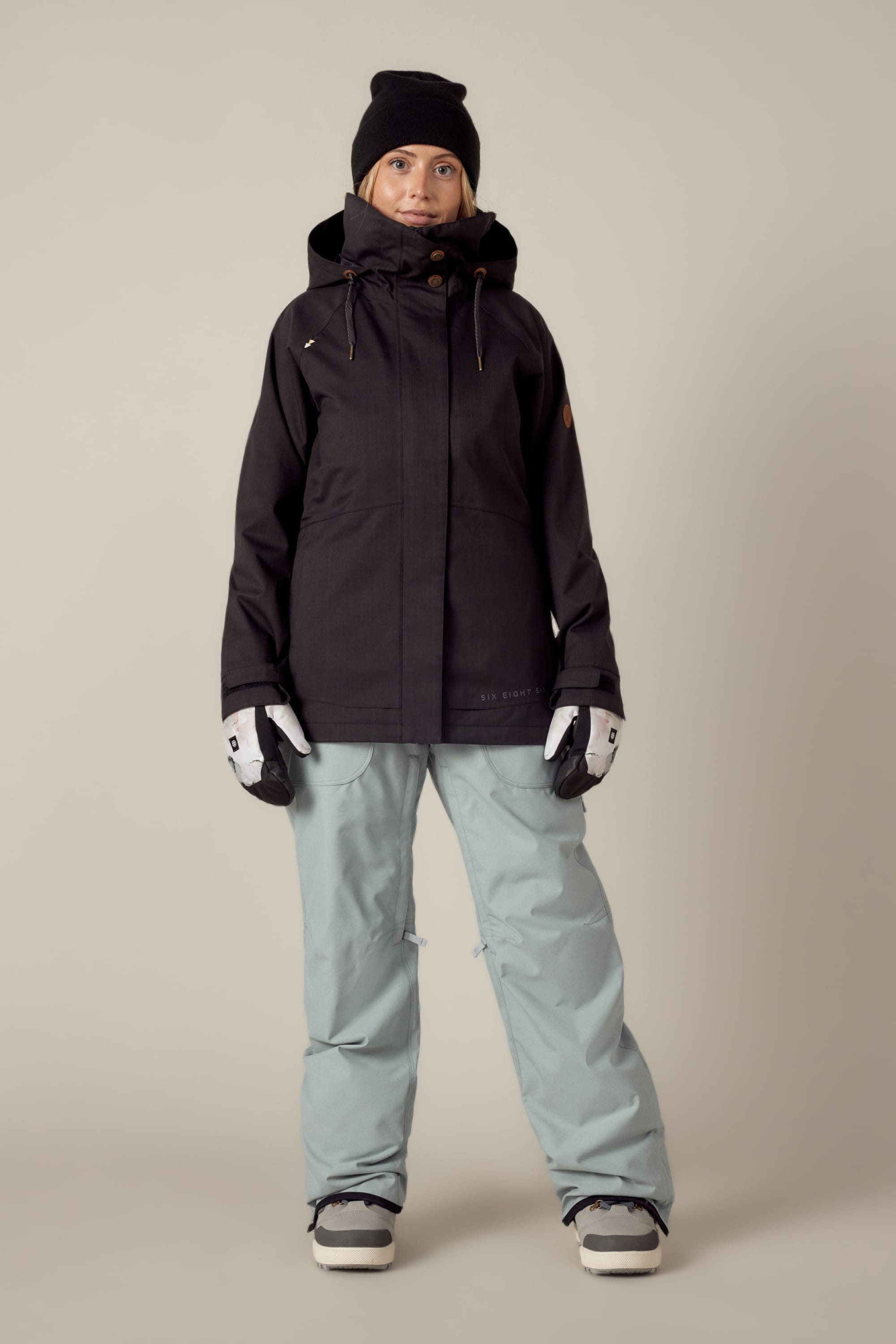 A person stands facing forward, wearing a 686 Womens Smarty 3-In-1 Spellbound Jacket in black, light blue snow pants, a black beanie, white gloves, and snow boots against a plain beige background.
