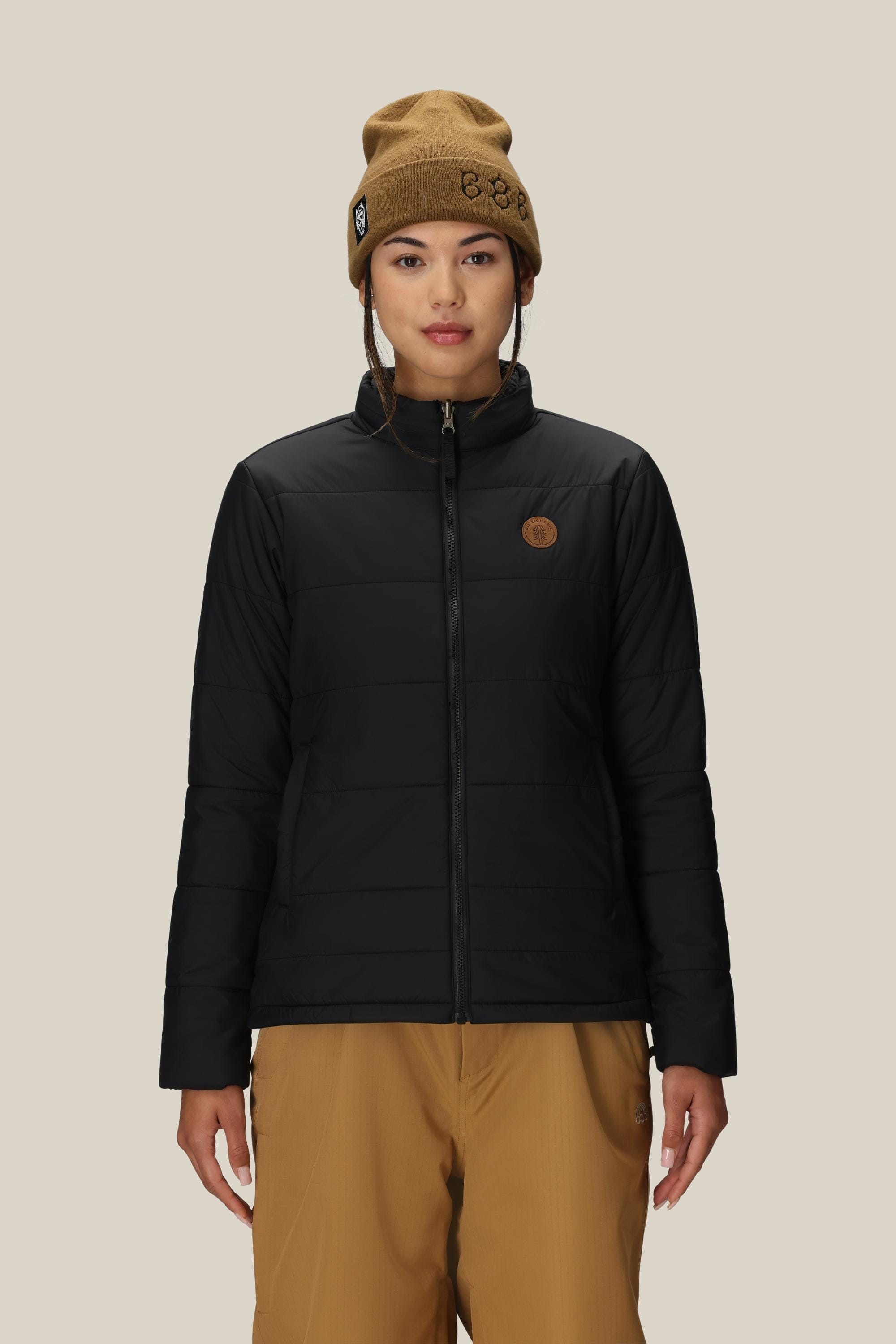 A woman wearing the 686 Womens Smarty 3-In-1 Spellbound Jacket in black, with tan pants and a tan beanie, stands facing forward against a plain light beige background.