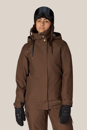 A woman dressed in the 686 Women’s SMARTY® 3-In-1 Spellbound Jacket stands against a plain light background, paired with matching pants, black gloves, and black ski goggles.