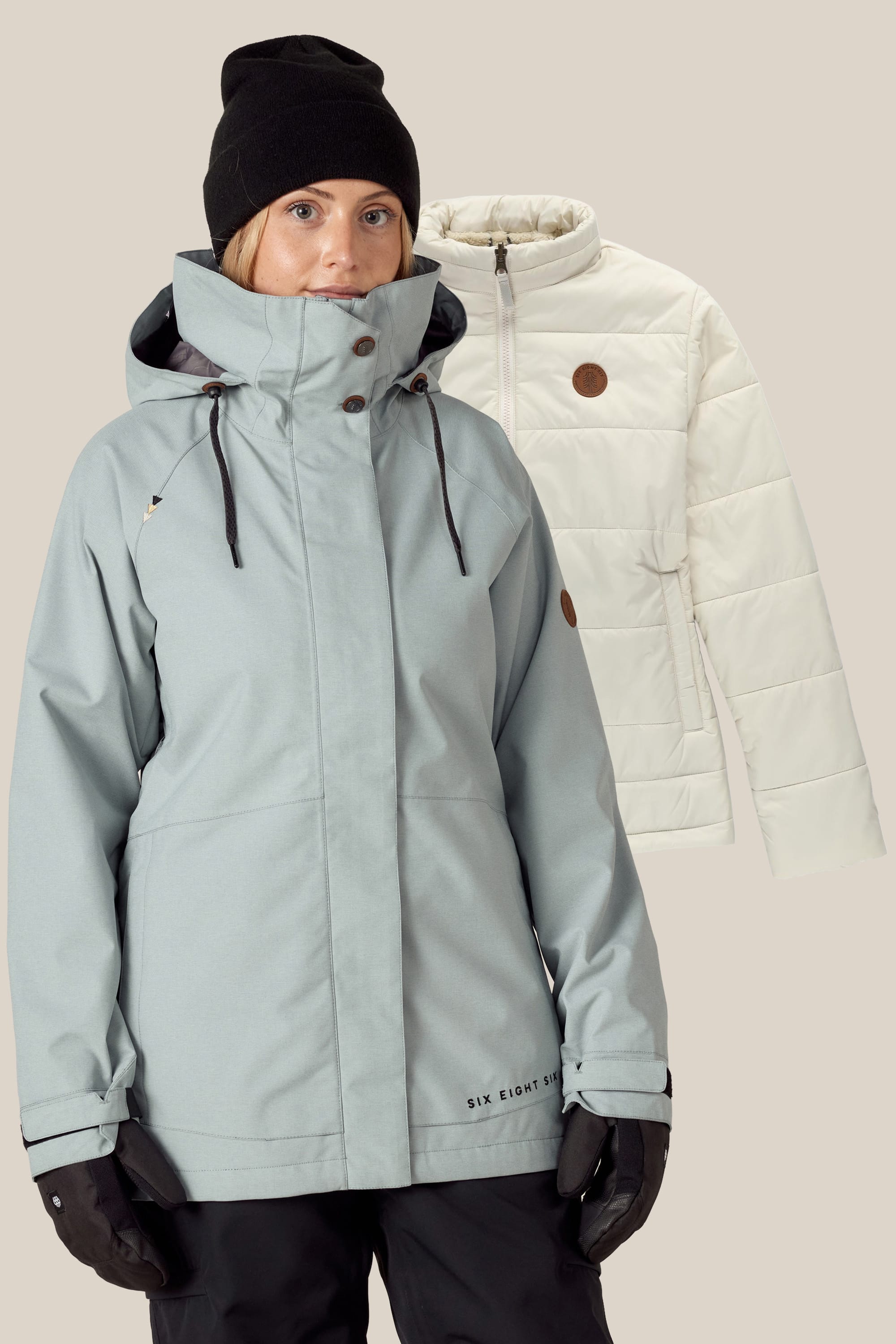 A woman in a light gray 686 Womens Smarty 3-In-1 Spellbound Jacket, black beanie, and gloves stands in front of a cream quilted jacket displayed in the background.