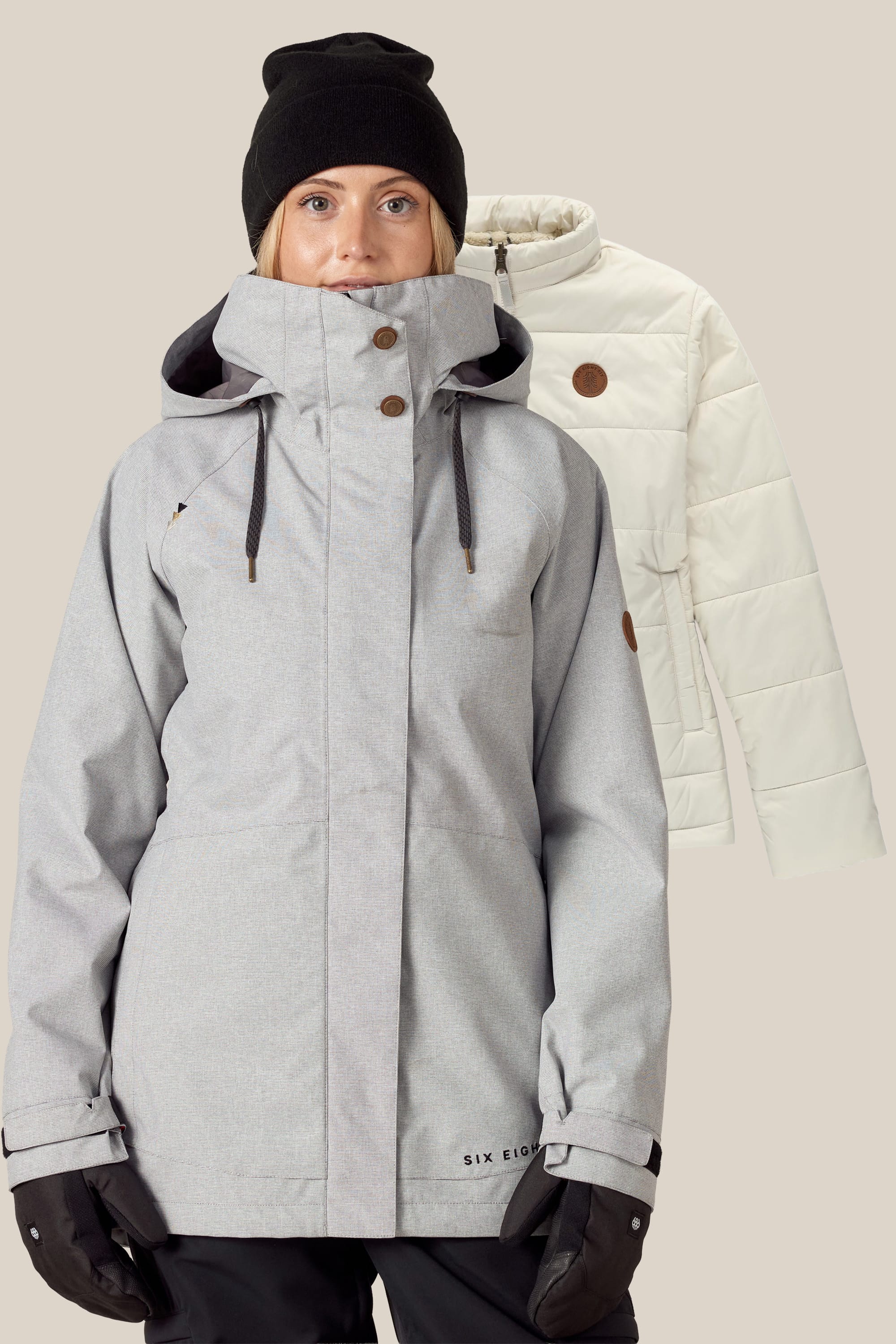 A woman wearing a light gray 686 Womens Smarty 3-In-1 Spellbound Jacket with a hood, black gloves, and a black beanie stands in front of a cream-colored quilted jacket in the background.