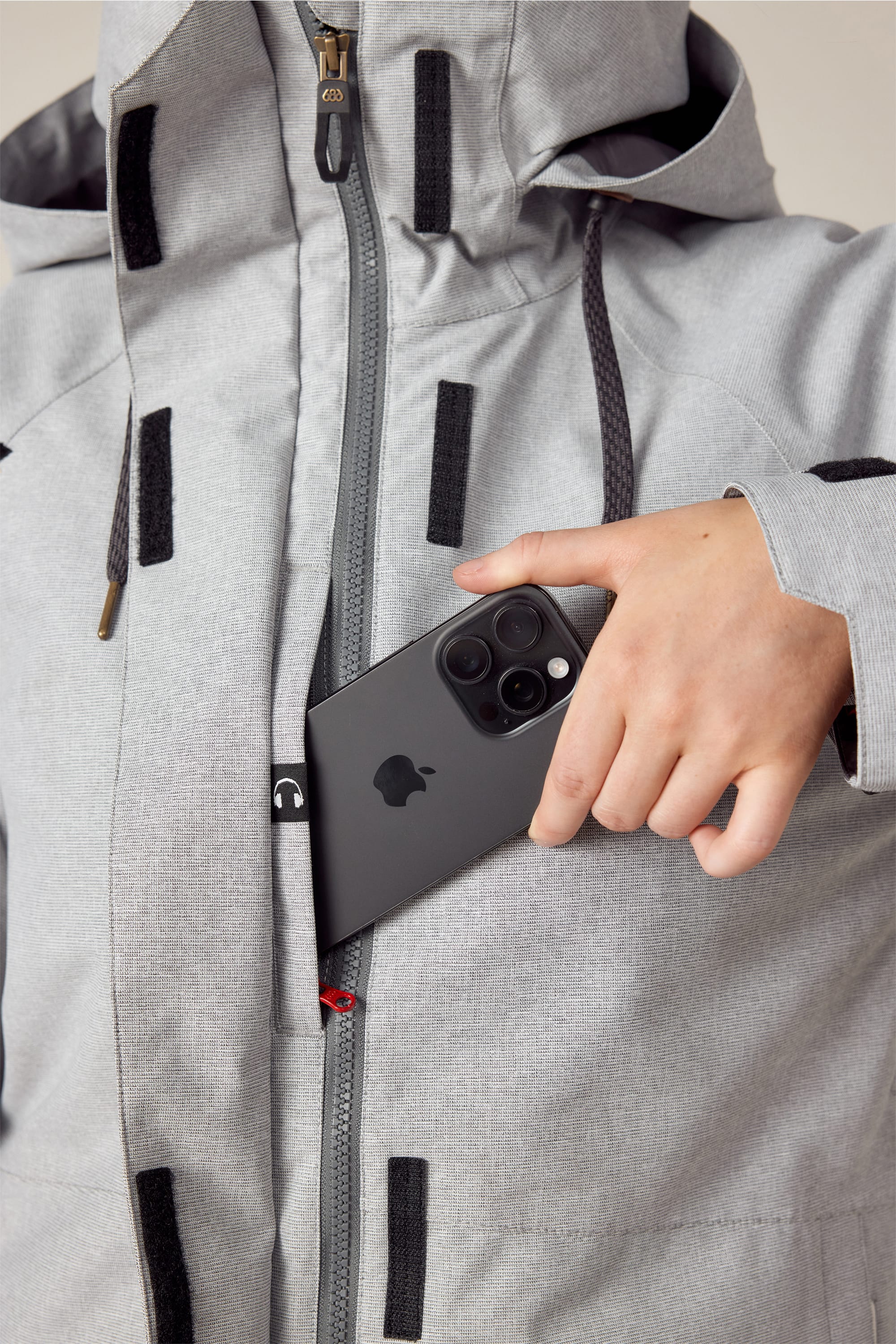 A woman in a light gray 686 Womens Smarty 3-In-1 Spellbound Jacket puts a black smartphone with three camera lenses into the vertically zippered chest pocket.