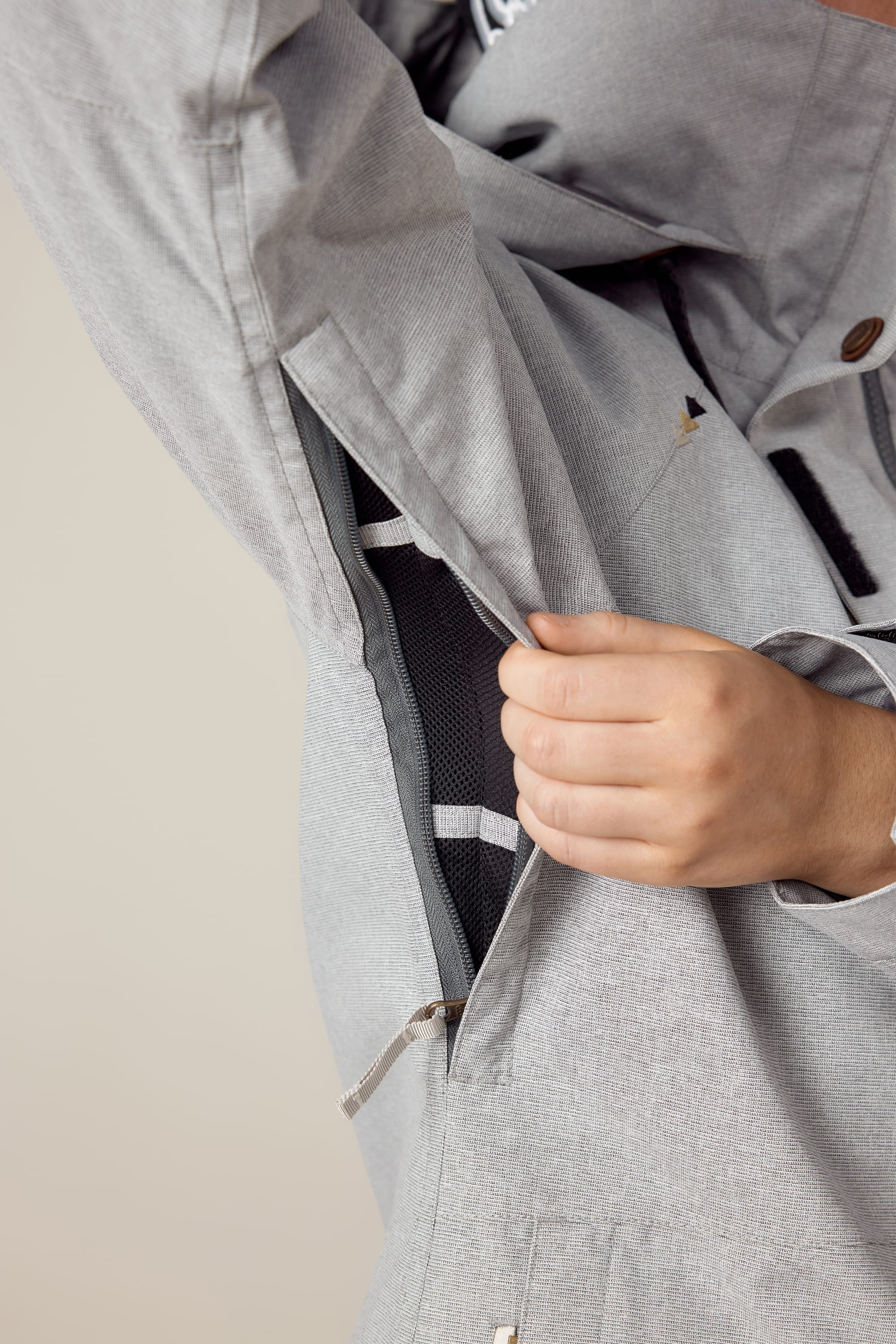A person opens a zippered underarm vent of the 686 Womens Smarty 3-In-1 Spellbound Jacket, revealing black mesh lining inside the light gray polyester outer shell.
