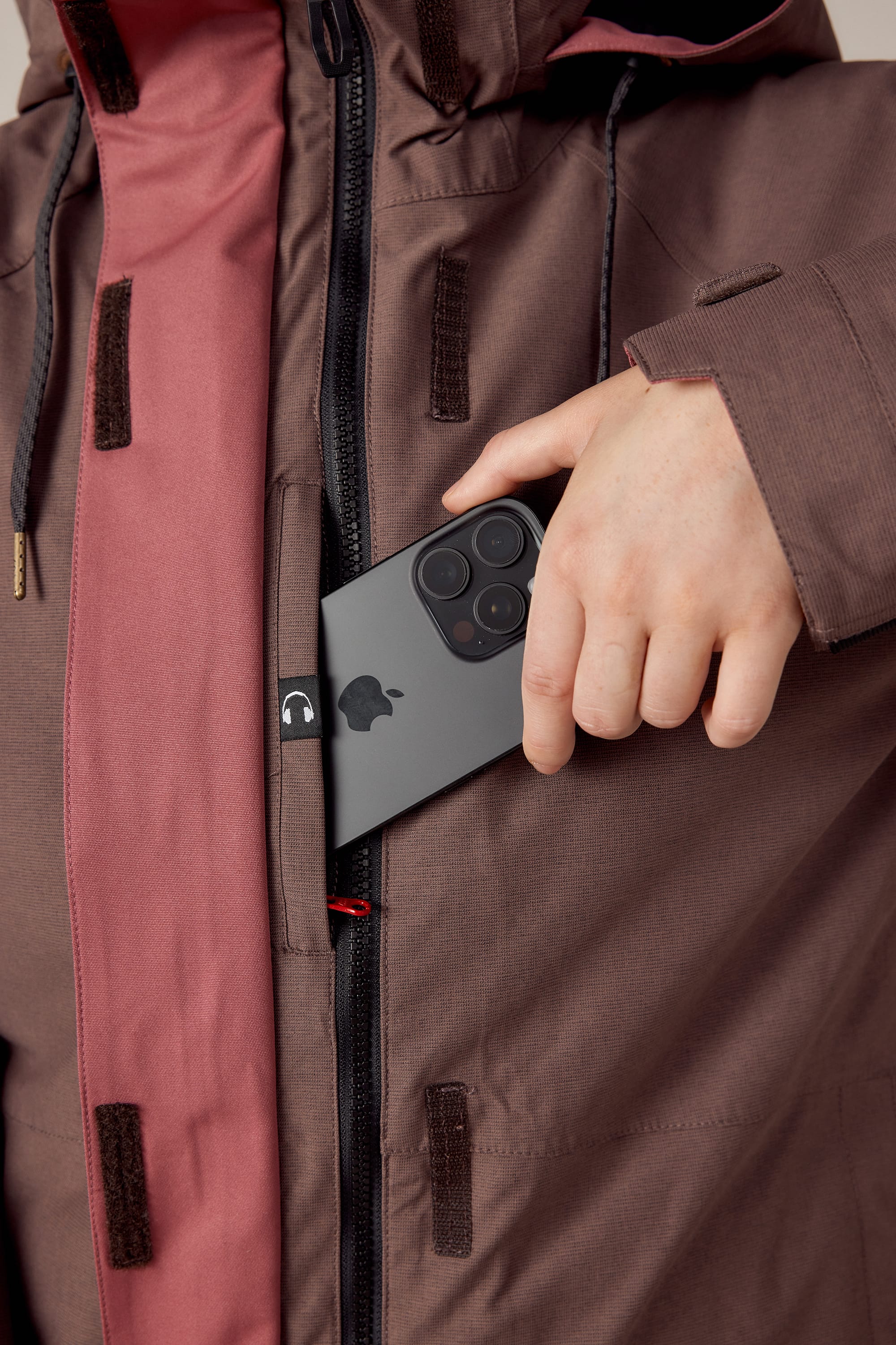 A woman in a 686 Womens Smarty 3-In-1 Spellbound Jacket (brown and pink polyester, Velcro fastenings) slips a triple-camera black iPhone into an inner pocket of the unzipped, stylish jacket by 686.