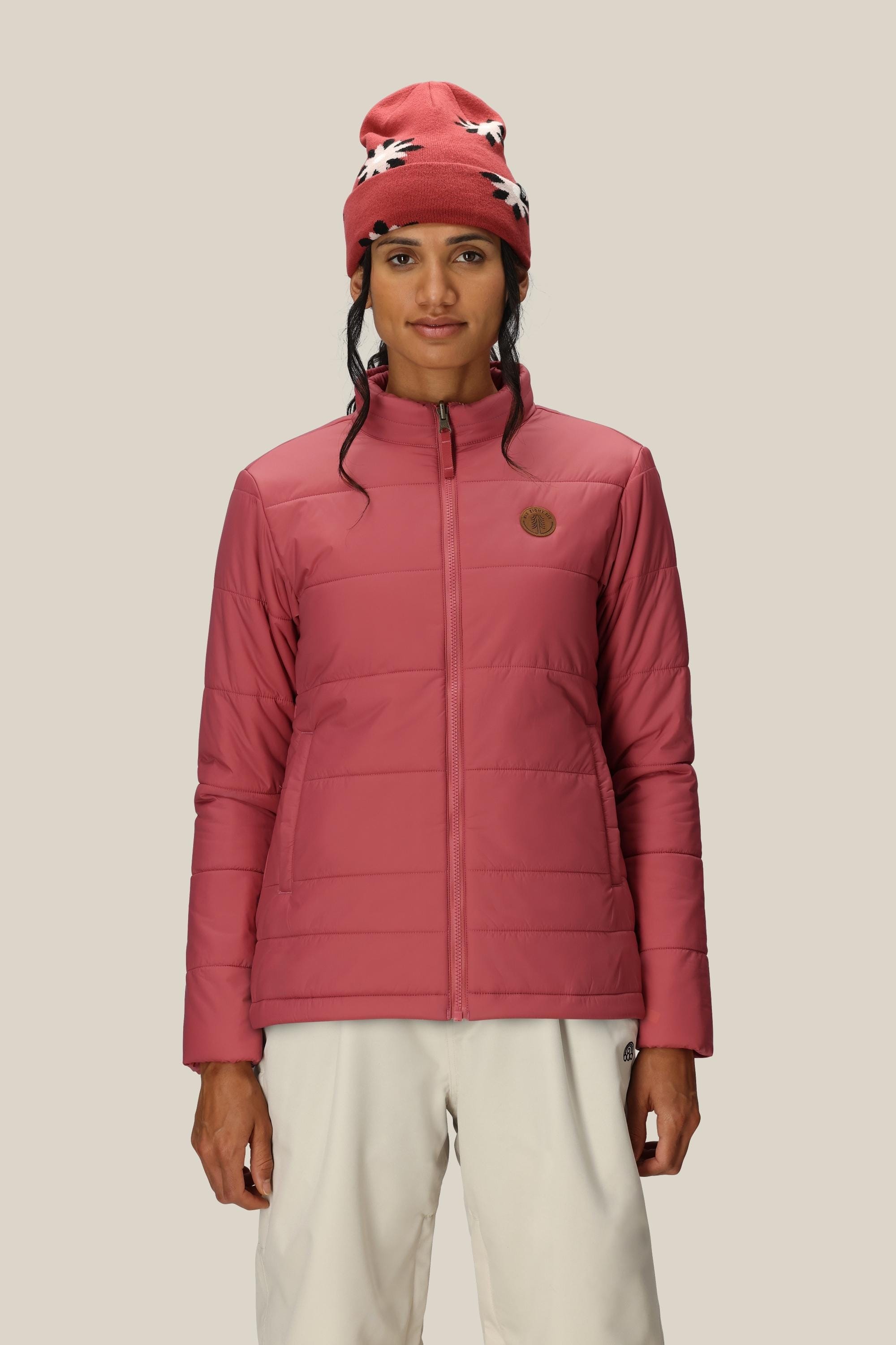 A woman wearing the 686 Womens Smarty 3-In-1 Spellbound Jacket in coral pink and a knit beanie with a white and black pattern stands against a plain beige background.