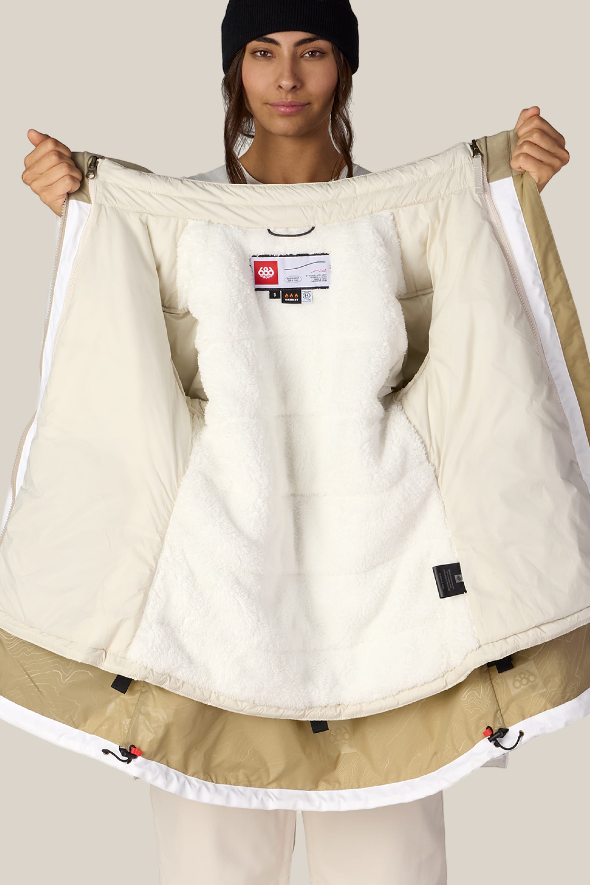 A person wearing a black beanie holds open the 686 Women’s SMARTY® 3-In-1 Spellbound Jacket, revealing its soft, plush white lining and interior labels. The background is plain and neutral.