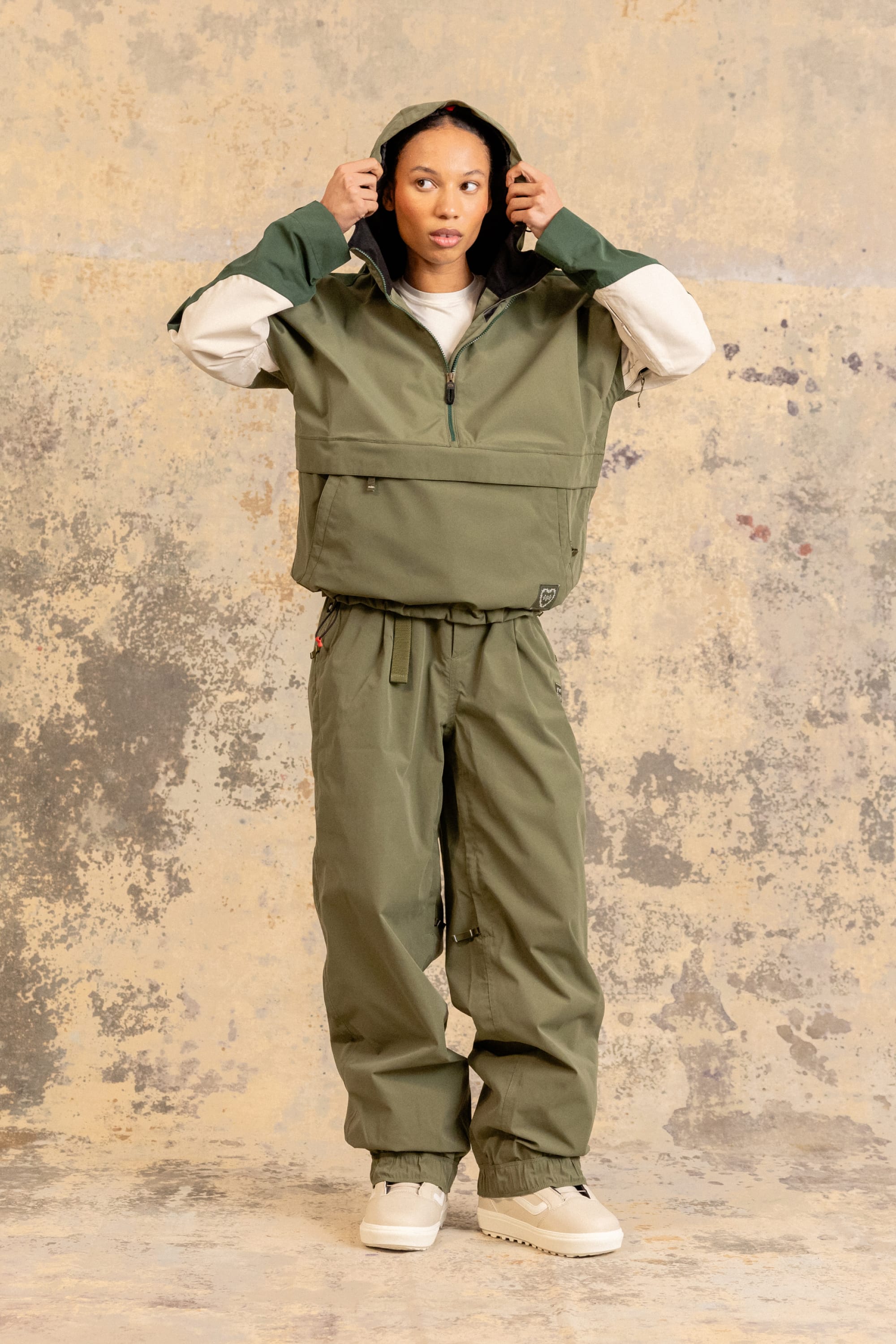 A person stands against a textured wall, wearing the 686 Women’s Outline™ Shell Anorak and matching pants with white sneakers. They hold the hood with both hands and look forward.