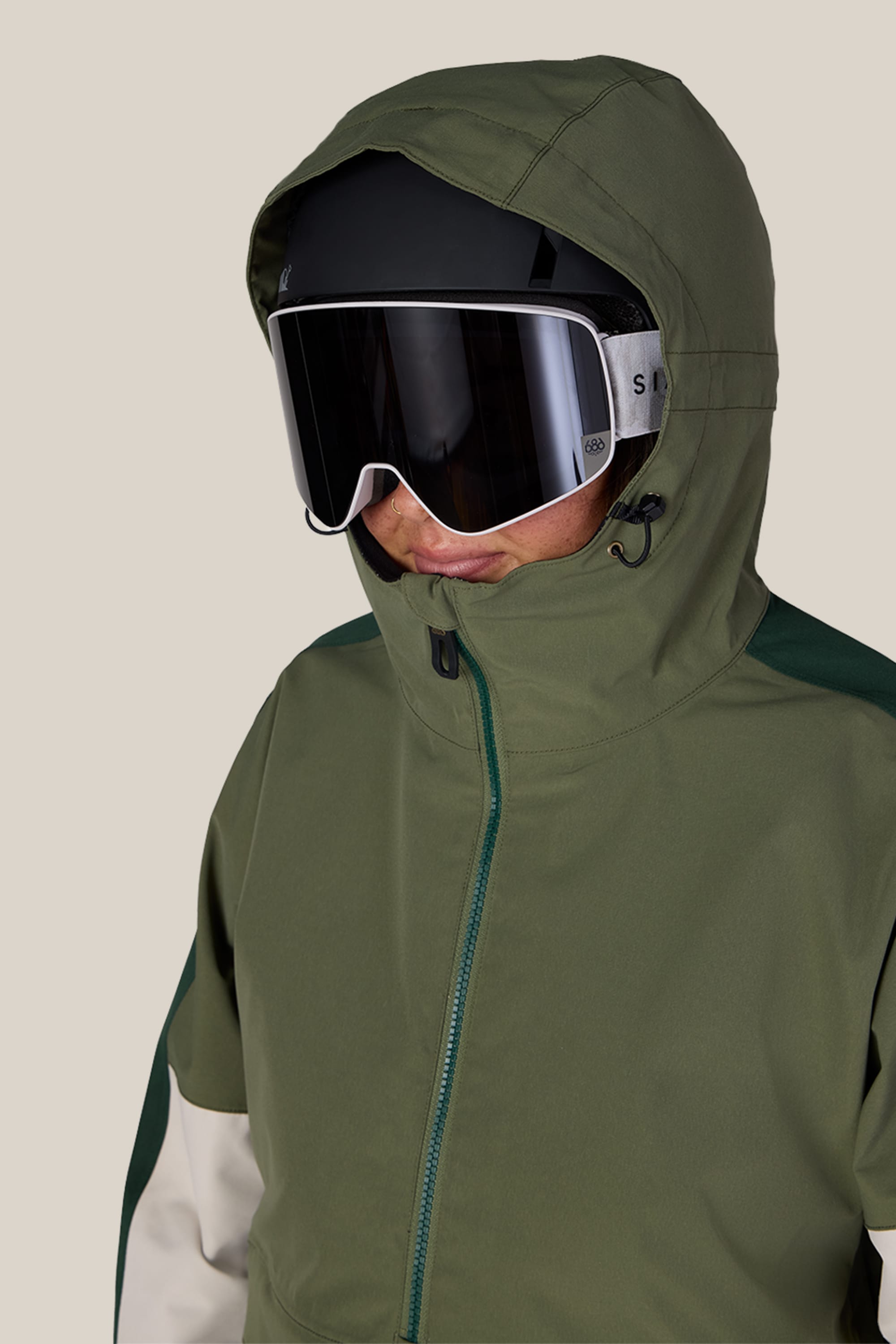 A woman models the 686 Women’s Outline™ Shell Anorak in green, paired with black ski goggles, as she faces slightly to the side against a plain beige background.