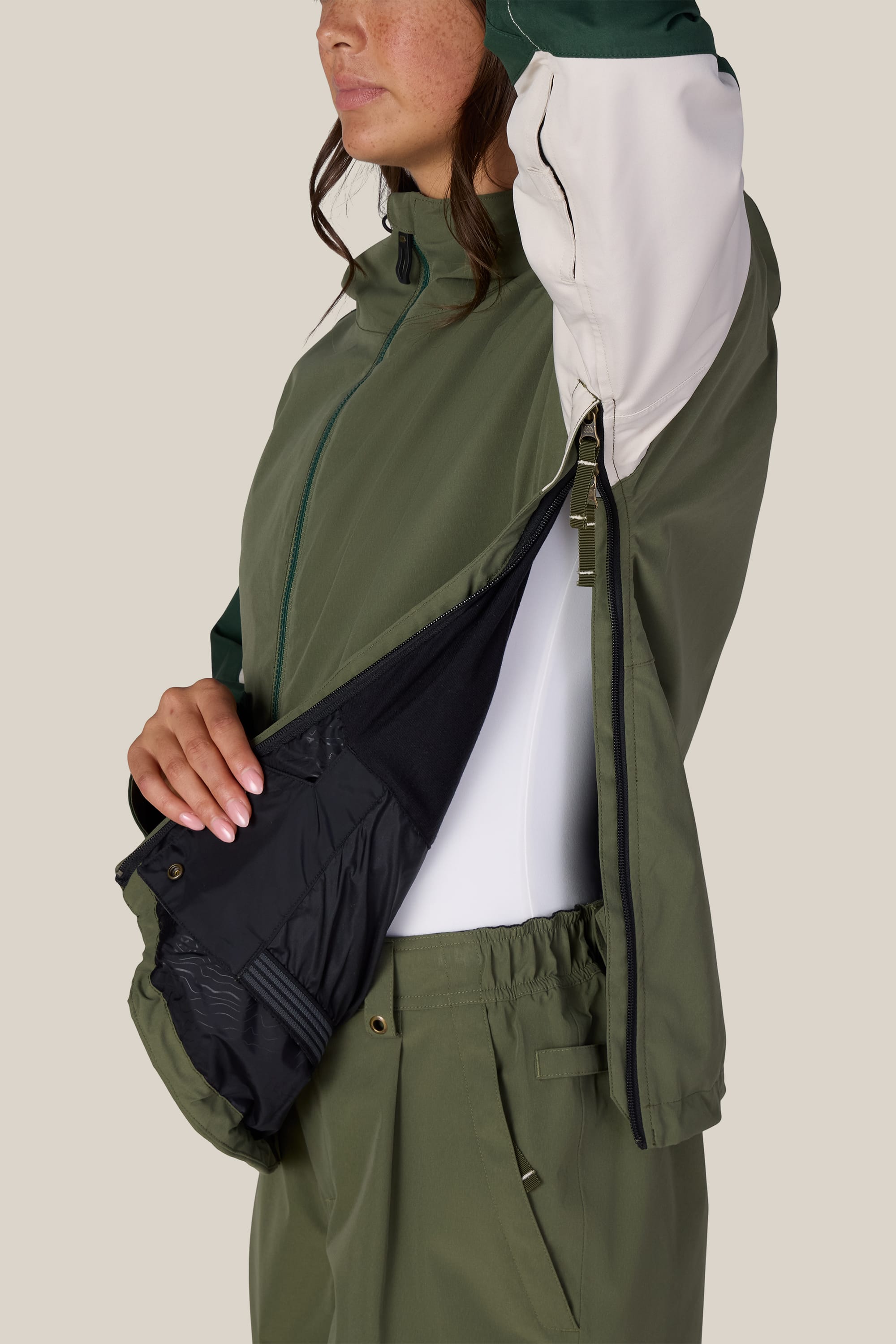 A person models the 686 Women’s Outline™ Shell Anorak in green and white, demonstrating the side zipper and revealing the jacket’s ventilated black mesh lining. The individual’s face is partially visible, with one arm raised.