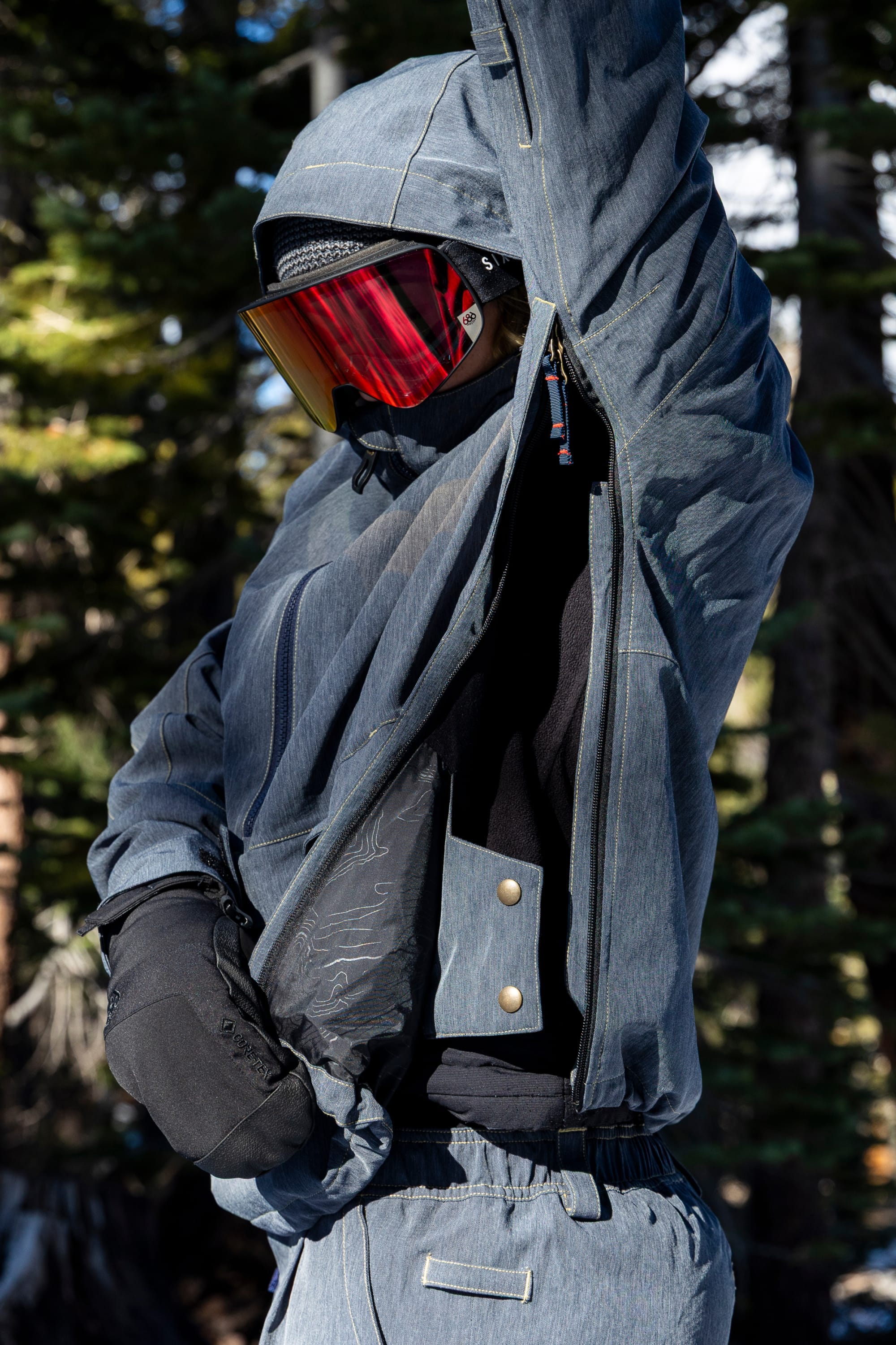 Clad in the 686 Women’s Outline™ Shell Anorak and red goggles, a person lifts their arm to display the jacket’s large zippered vent amid a snowy forest backdrop.