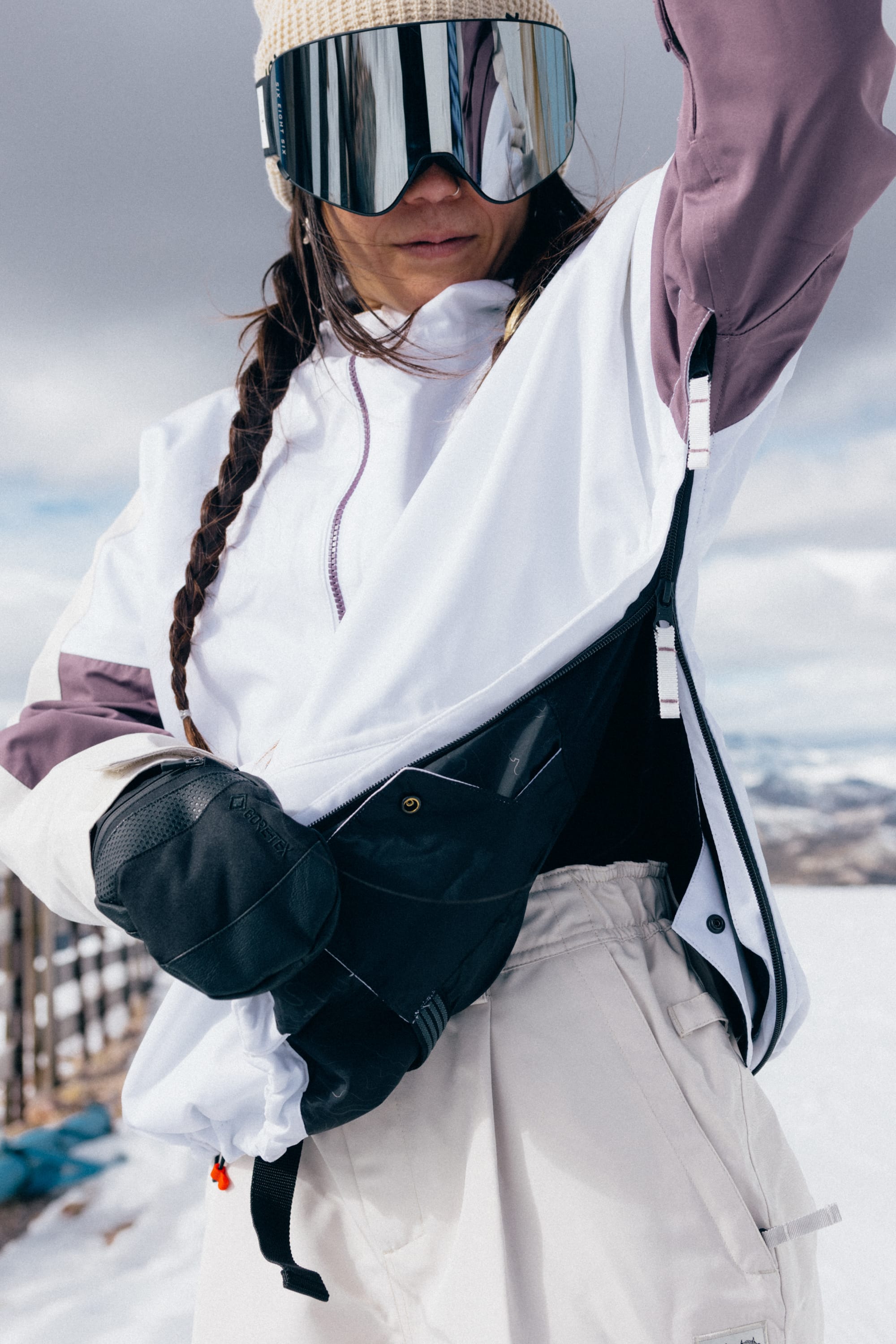A person in winter gear with braided hair and ski goggles stands outdoors in the snow, wearing the 686 Women’s Outline™ Shell Anorak in white and purple, beige pants, and black gloves, adjusting a black shoulder bag.