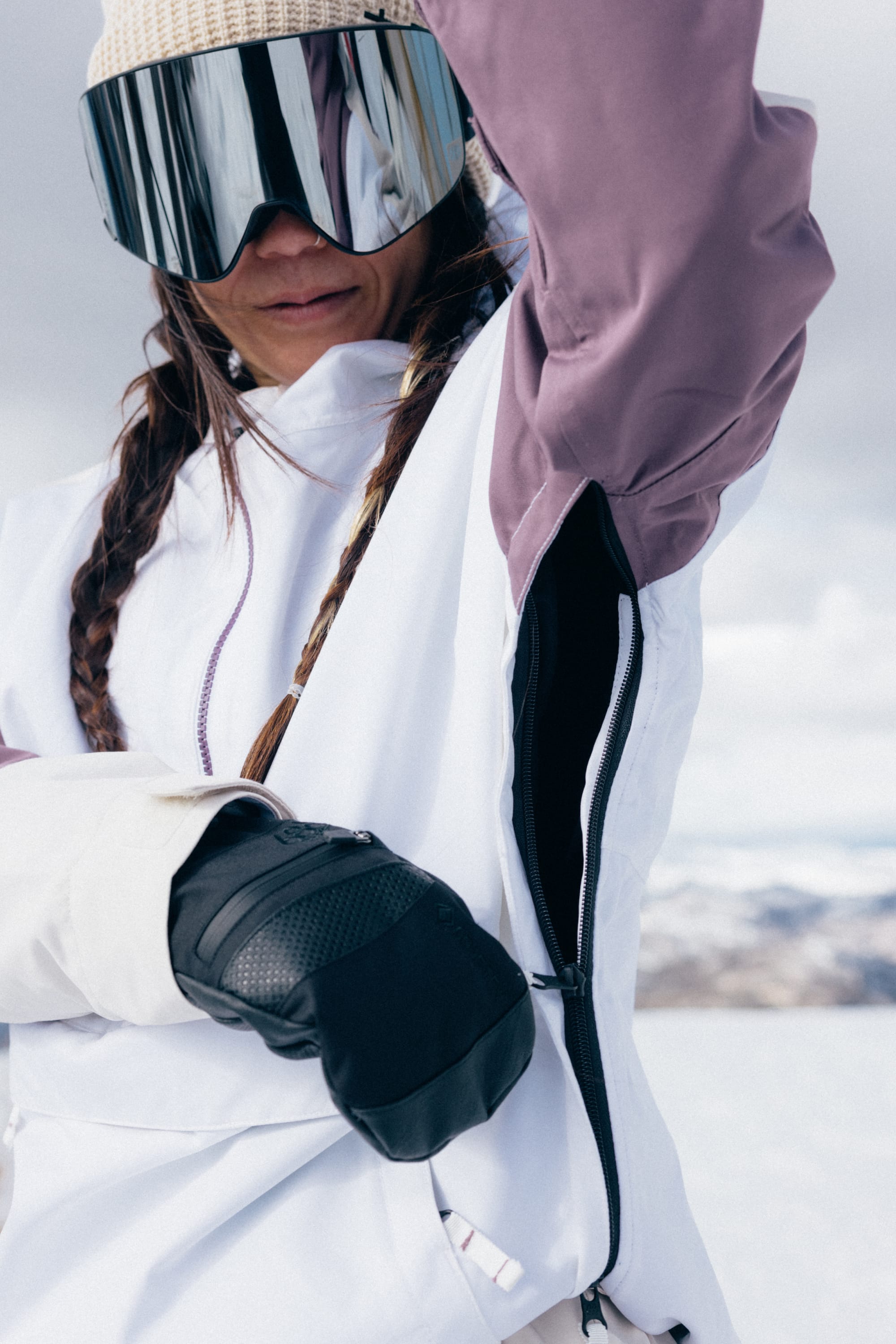 Wearing the 686 Women’s Outline™ Shell Anorak, a person in ski gear with tinted goggles, a knit hat, and braided hair adjusts the zipper on their white and purple jacket as snowy mountains rise in the background.