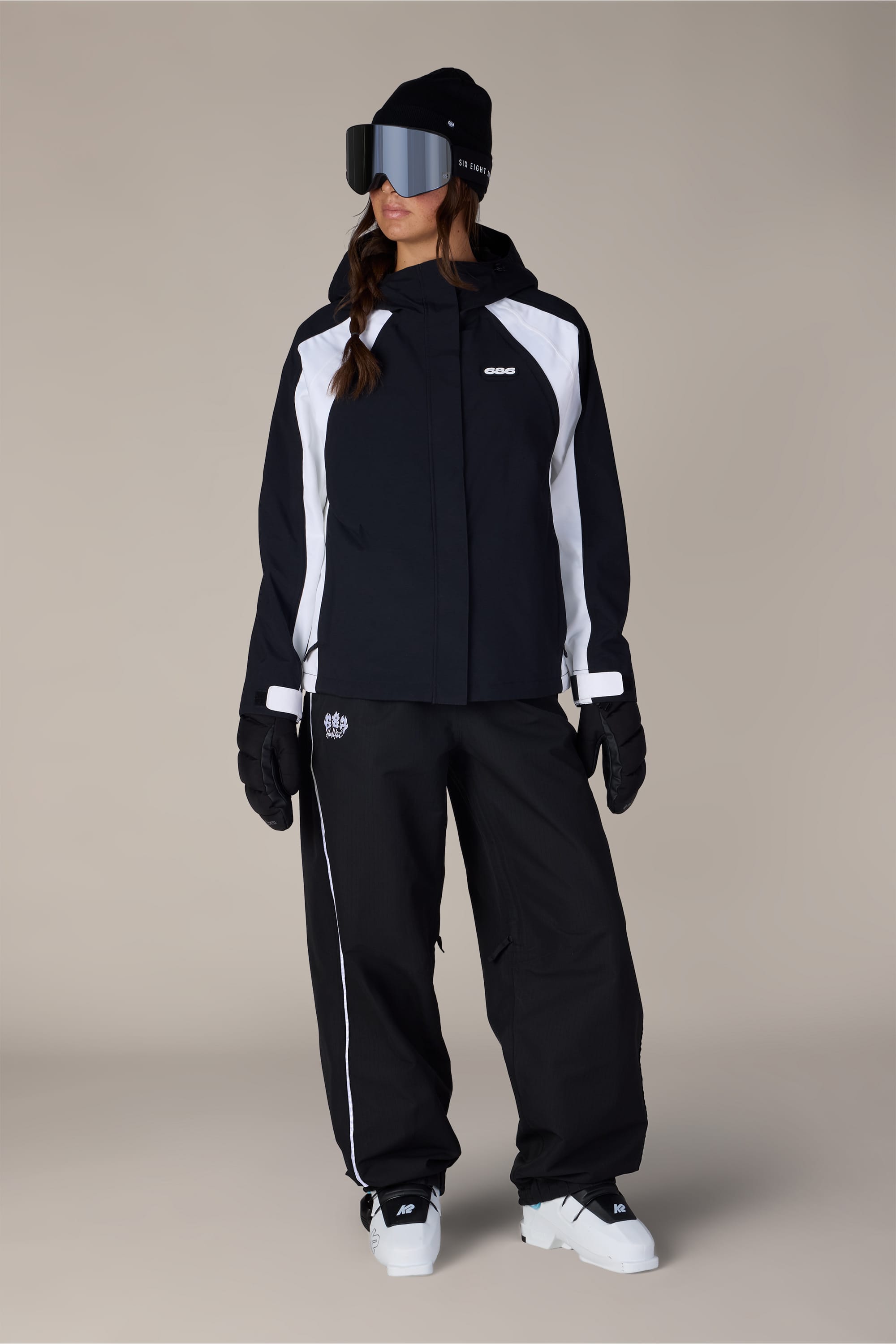 A person wearing the 686 Women’s Hologram™ Shell Jacket, along with gloves, white snow boots, a beanie, and large ski goggles, stands against a plain, light background.