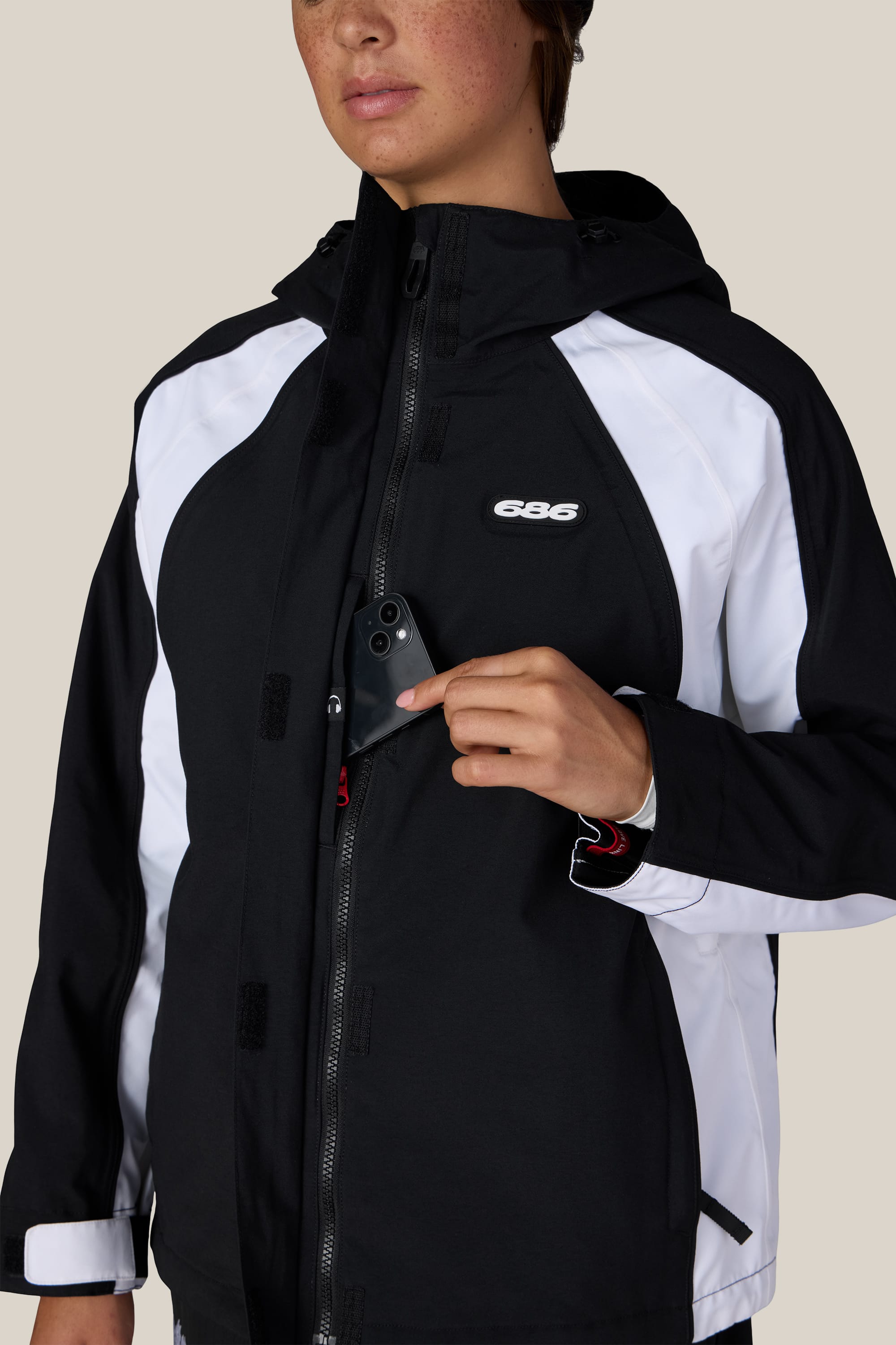 A person wearing the 686 Women’s Hologram™ Shell Jacket in black and white, featuring visible zippers, Velcro, and the 686 logo, places a phone into the chest pocket. The plain beige background contrasts with their partially visible face.