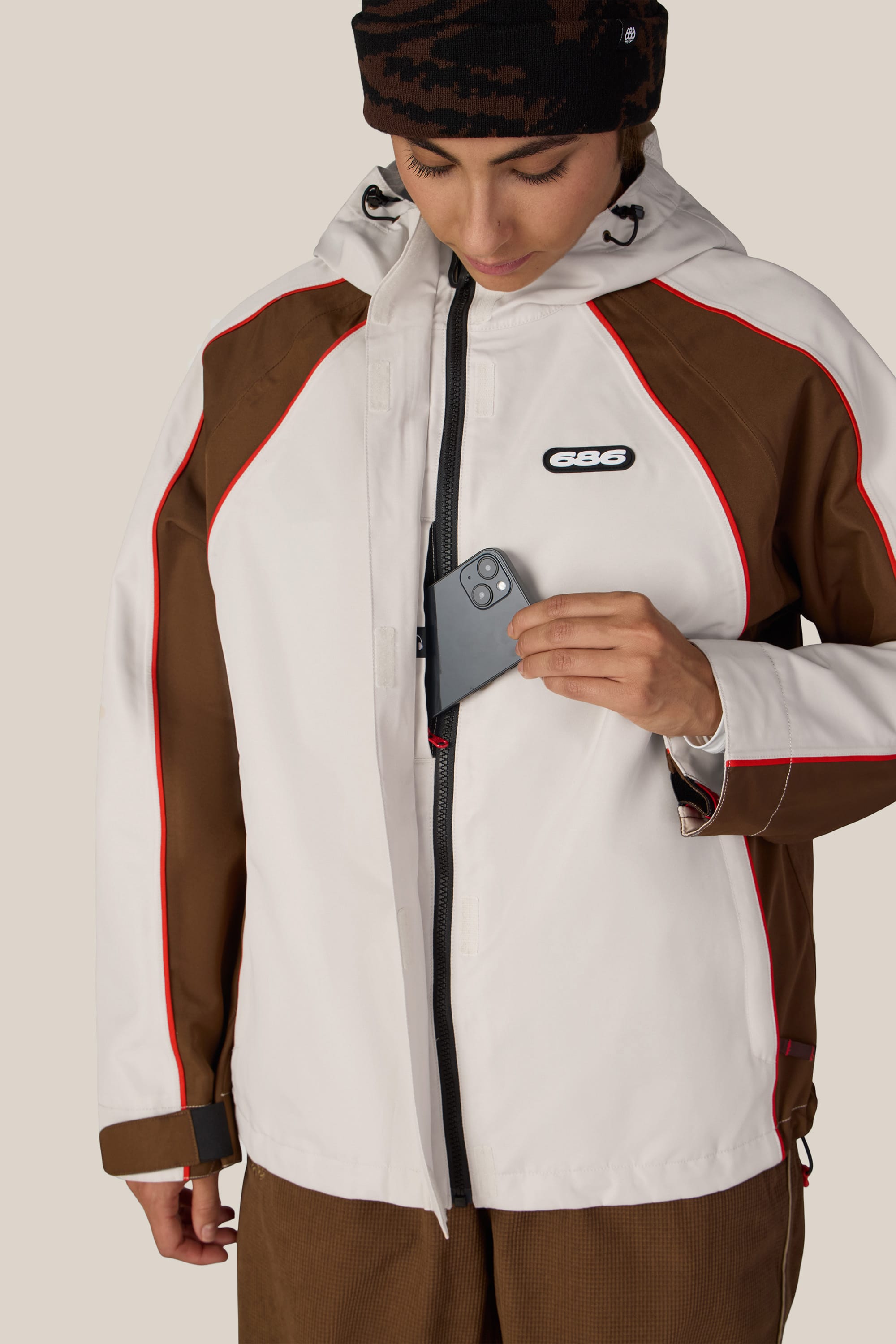 A woman wearing the 686 Women’s Hologram™ Shell Jacket with brown, white, and red accents places her smartphone into the inner chest pocket while looking down.