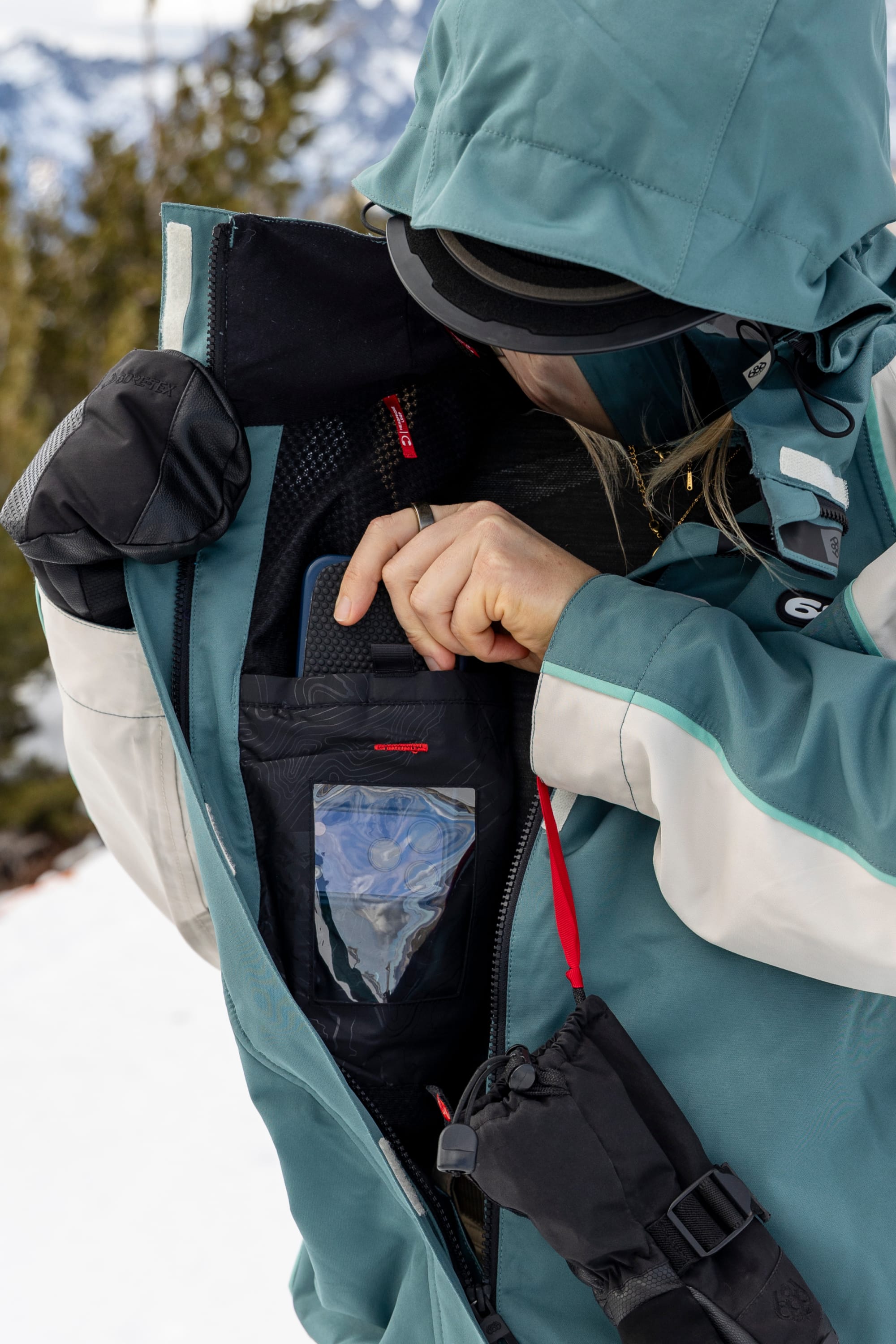 Wearing the 686 Women’s Hologram™ Shell Jacket, a person opens an inner pocket to store a smartphone, with snowy trees and mountains blurred in the background.