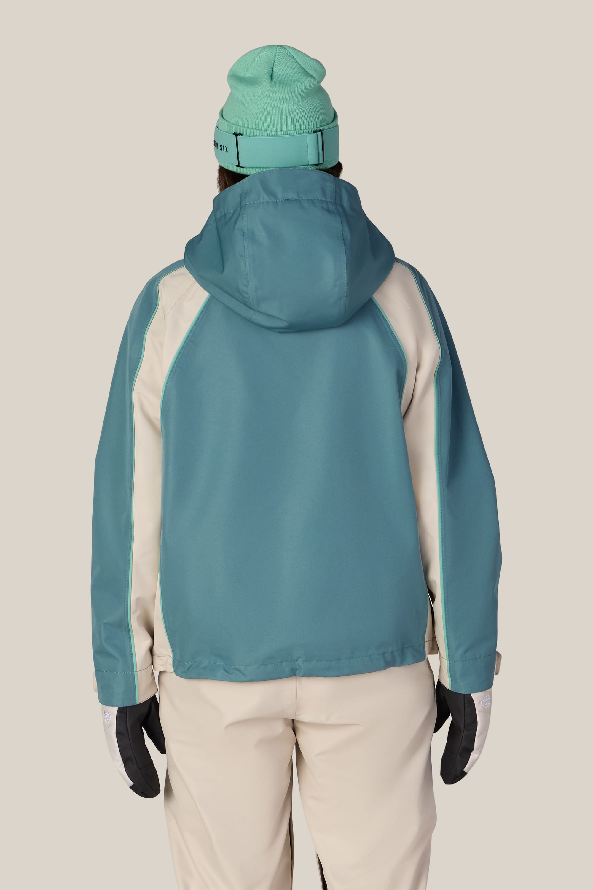 A person faces away, wearing the 686 Women’s Hologram™ Shell Jacket by 686 in teal with cream sleeves, paired with cream pants, black gloves, and a teal beanie against a plain light background.