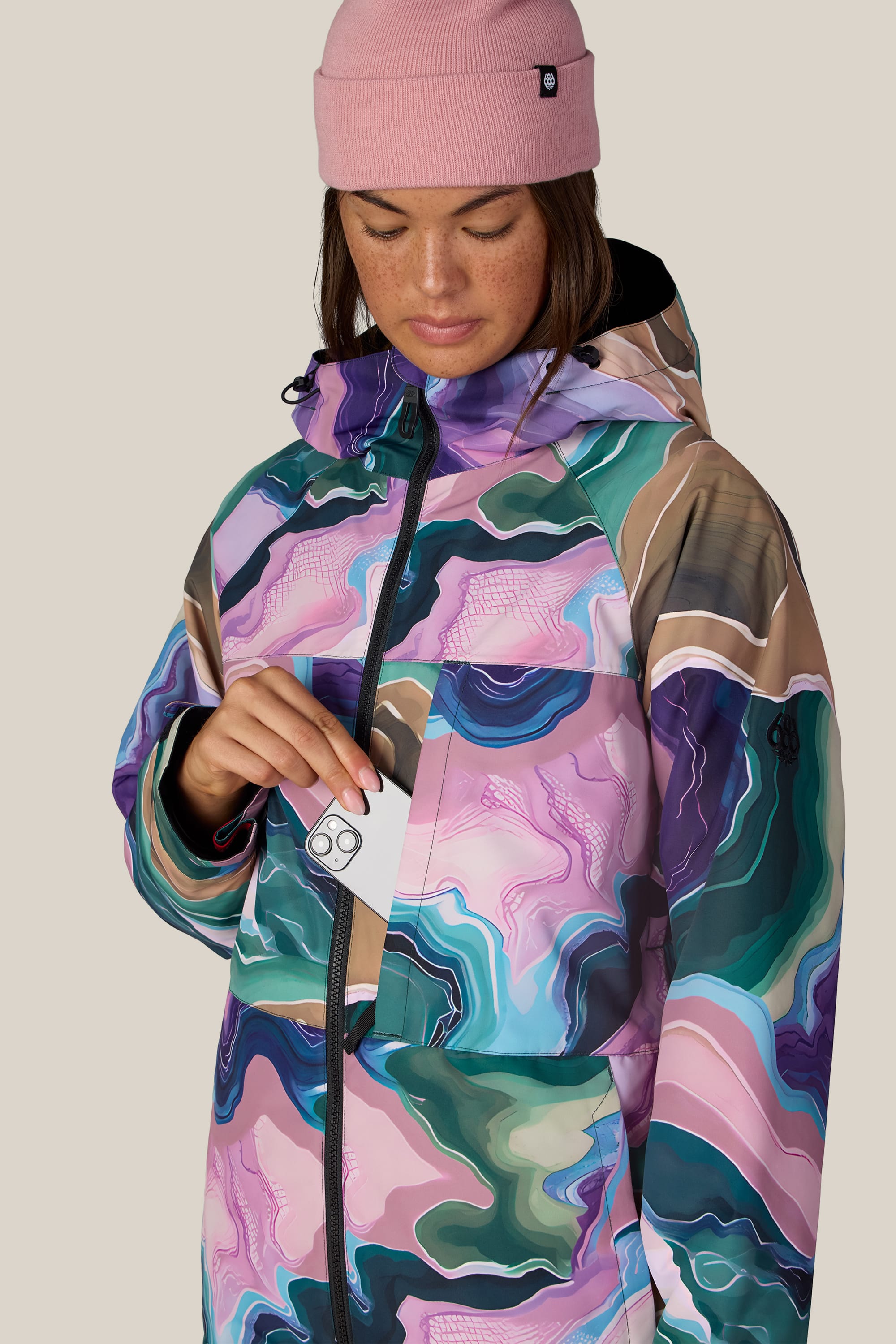 A woman wearing the 686 Women’s Athena™ Insulated Jacket and a pink beanie places a card into the front zipper pocket, looking down at the pocket.