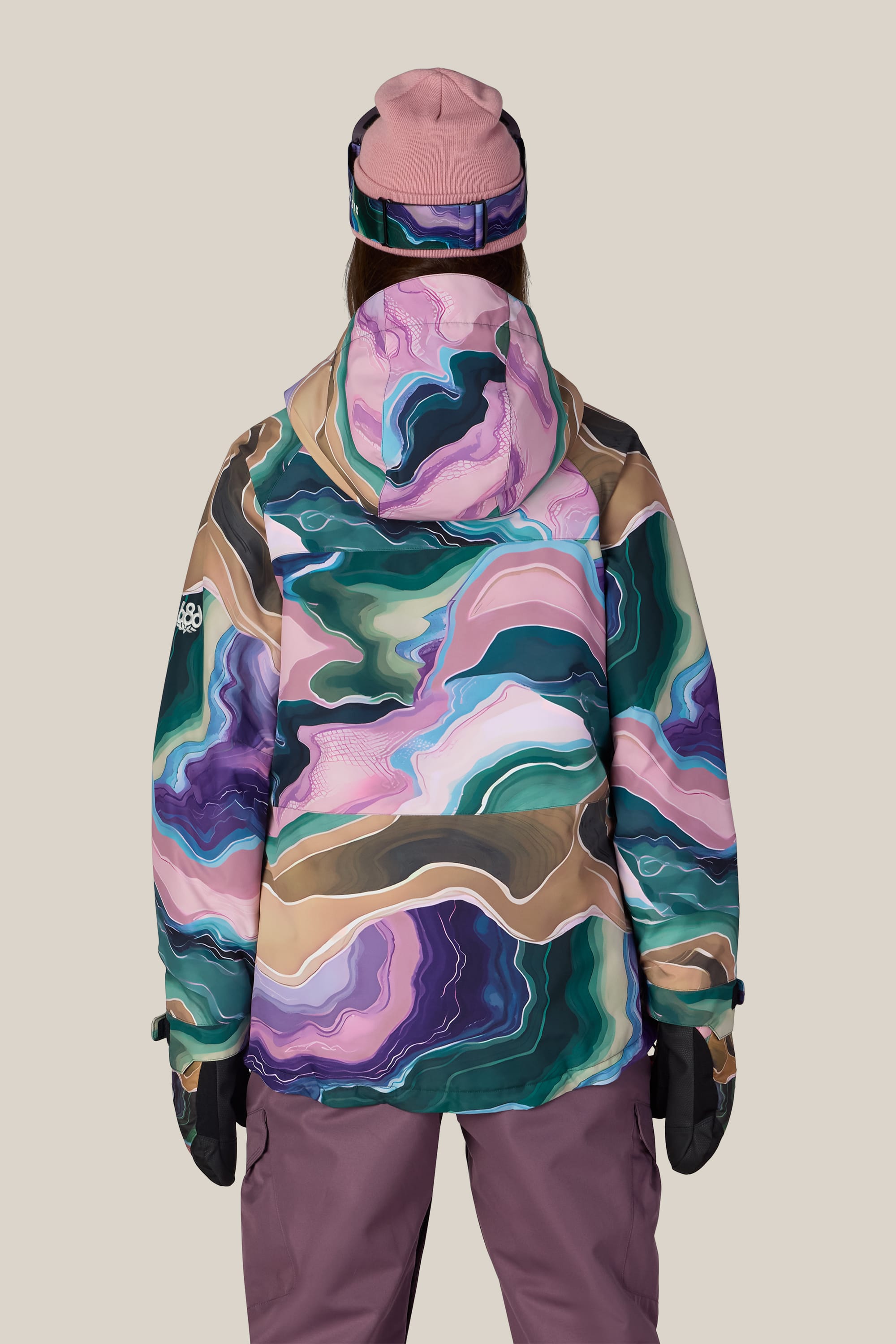 A person facing away is wearing the 686 Women’s Athena™ Insulated Jacket by 686, featuring a colorful abstract pattern in greens, purples, and pinks. They have on matching gloves and a pink beanie, set against a plain beige background.