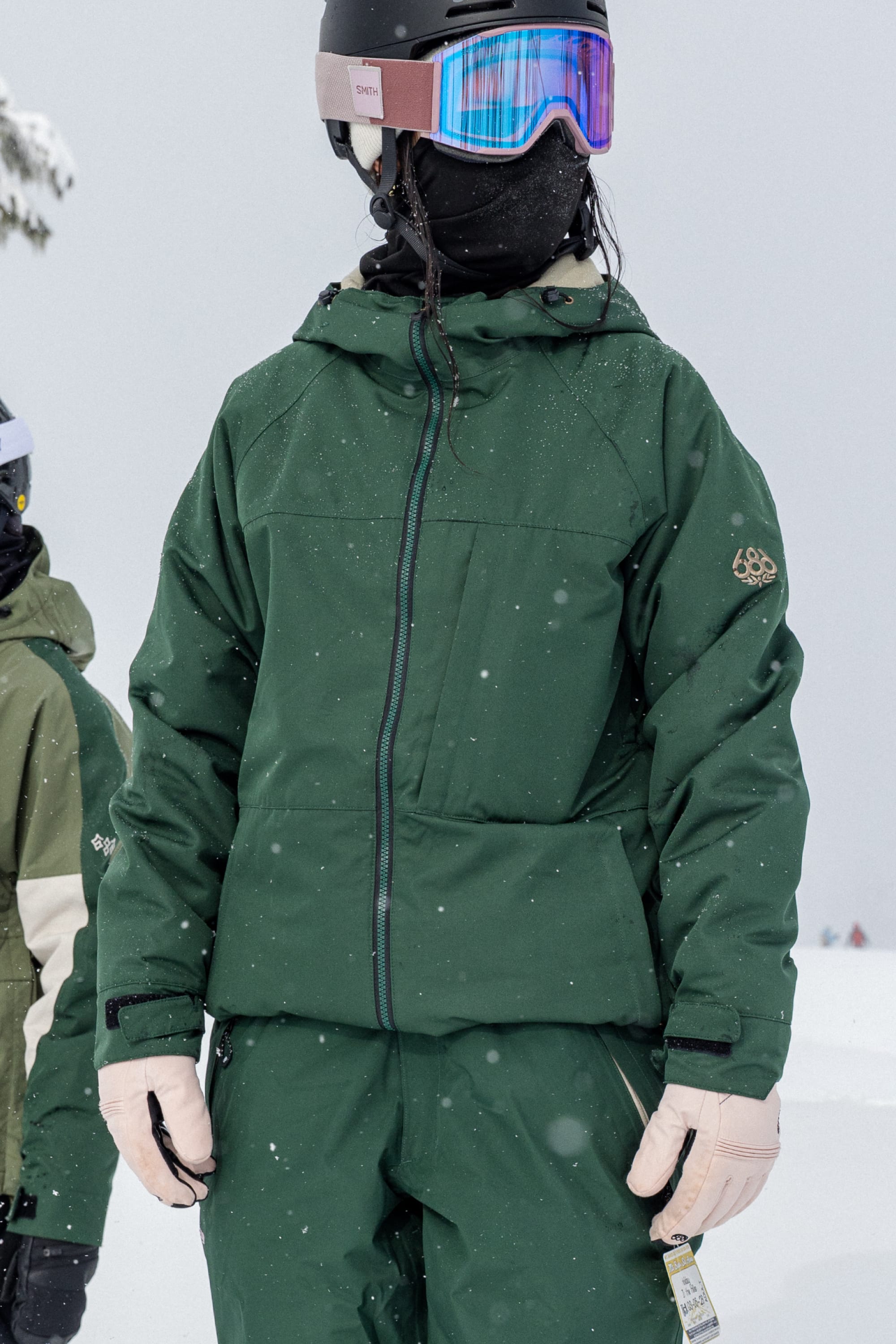 A person wearing the 686 Women’s Athena™ Insulated Jacket from 686, along with matching green pants, pink gloves, a helmet, and reflective goggles, stands in the snow as light snow falls. Another person is partially visible in the background.
