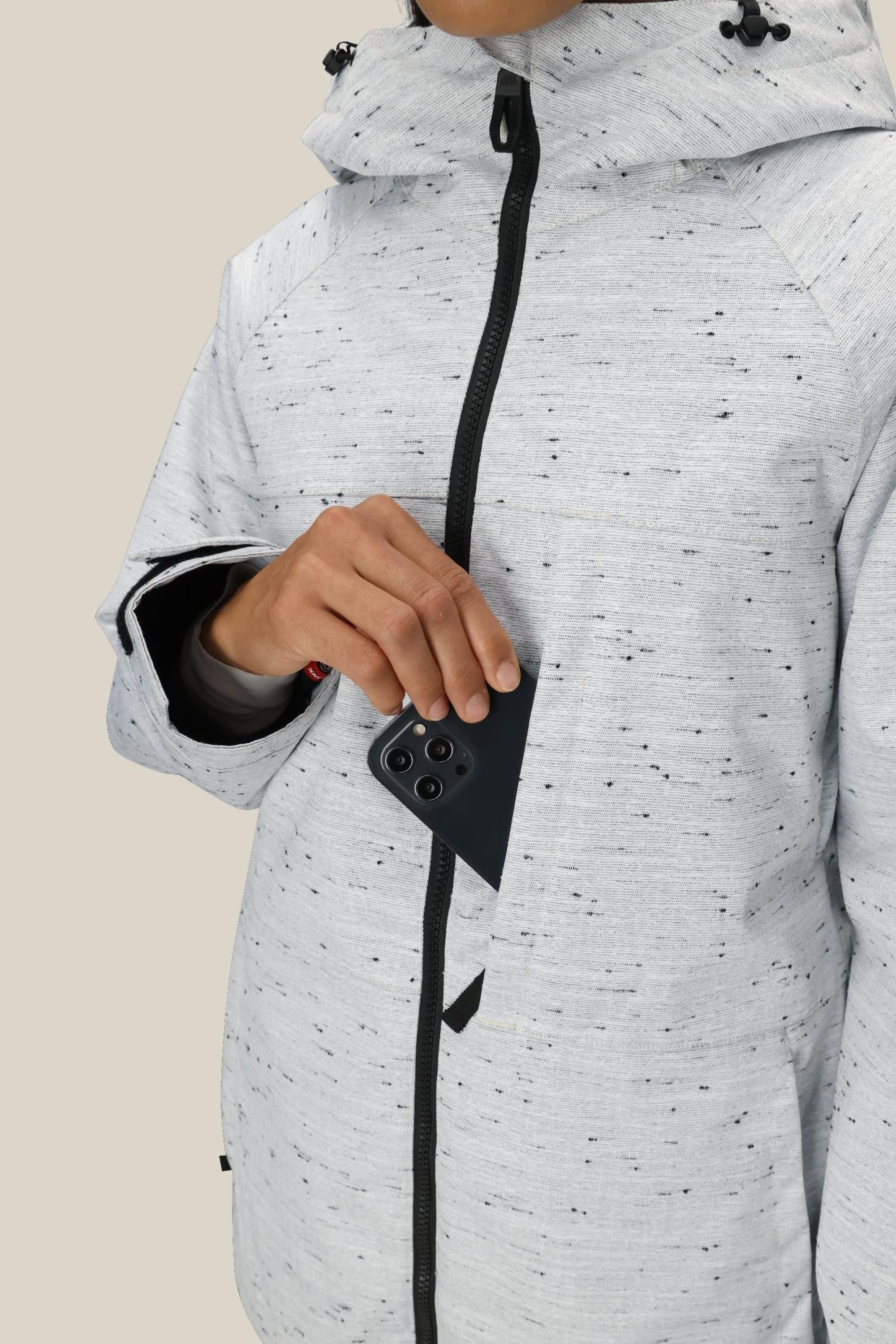 A person wearing the 686 Women’s Athena™ Insulated Jacket in light gray with black speckles is unzipping the chest pocket to place a black smartphone with three rear cameras inside; their face remains out of view.
