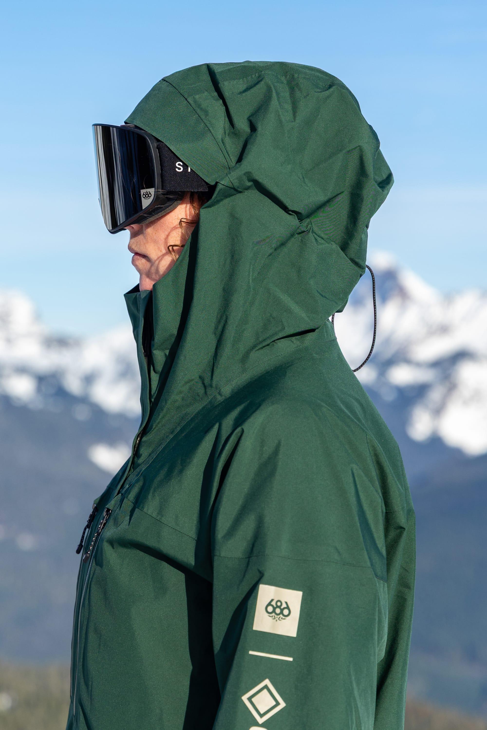 A person wearing the 686 Women’s GORE-TEX® Skyline Thermagraph® Jacket by 686, paired with black ski goggles, stands outdoors with snow-covered mountains and a blue sky in the background.