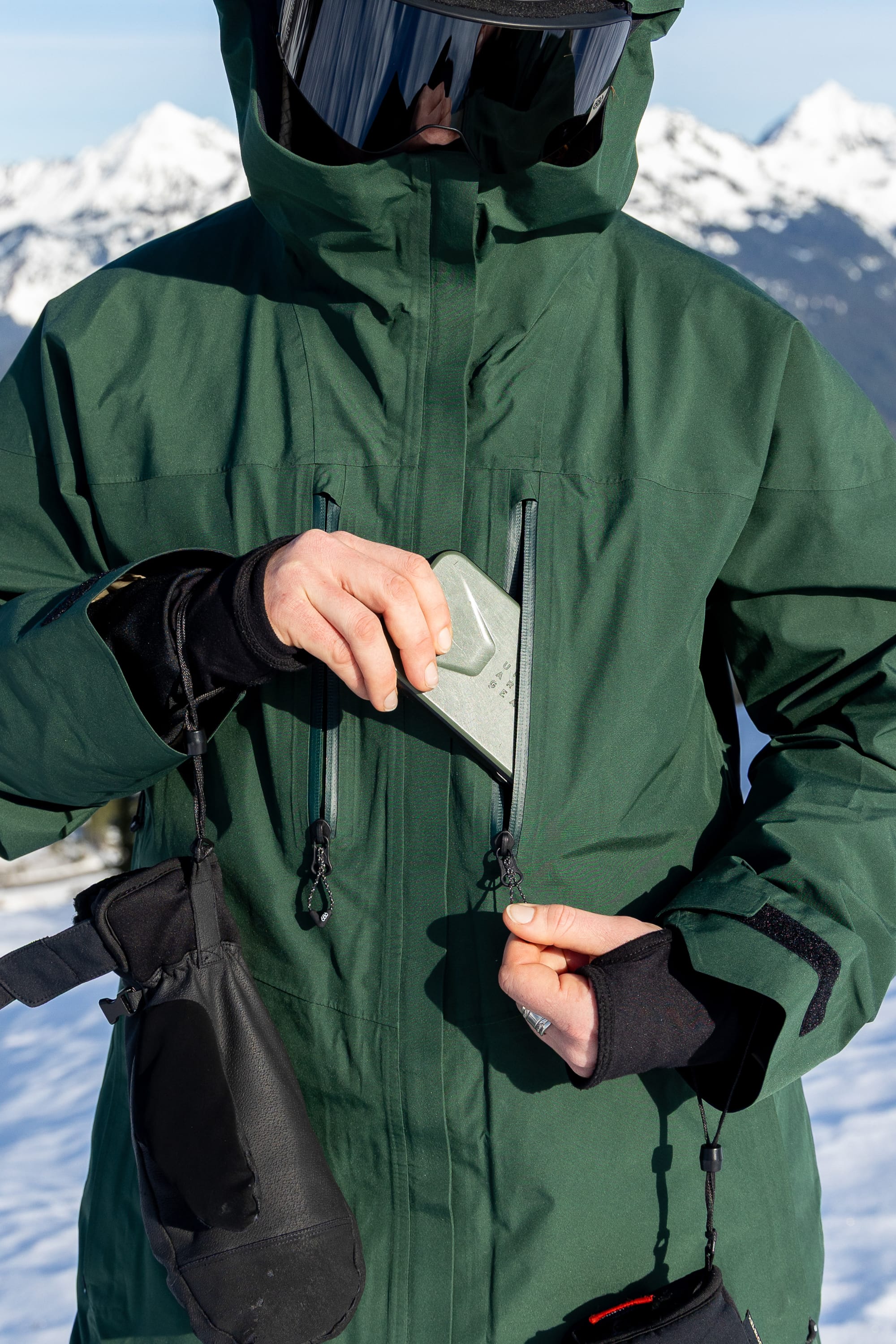 Wearing the 686 Women’s GORE-TEX® Skyline Thermagraph® Jacket, a person slides a silver smartphone into their chest pocket while standing in a snowy mountain landscape.