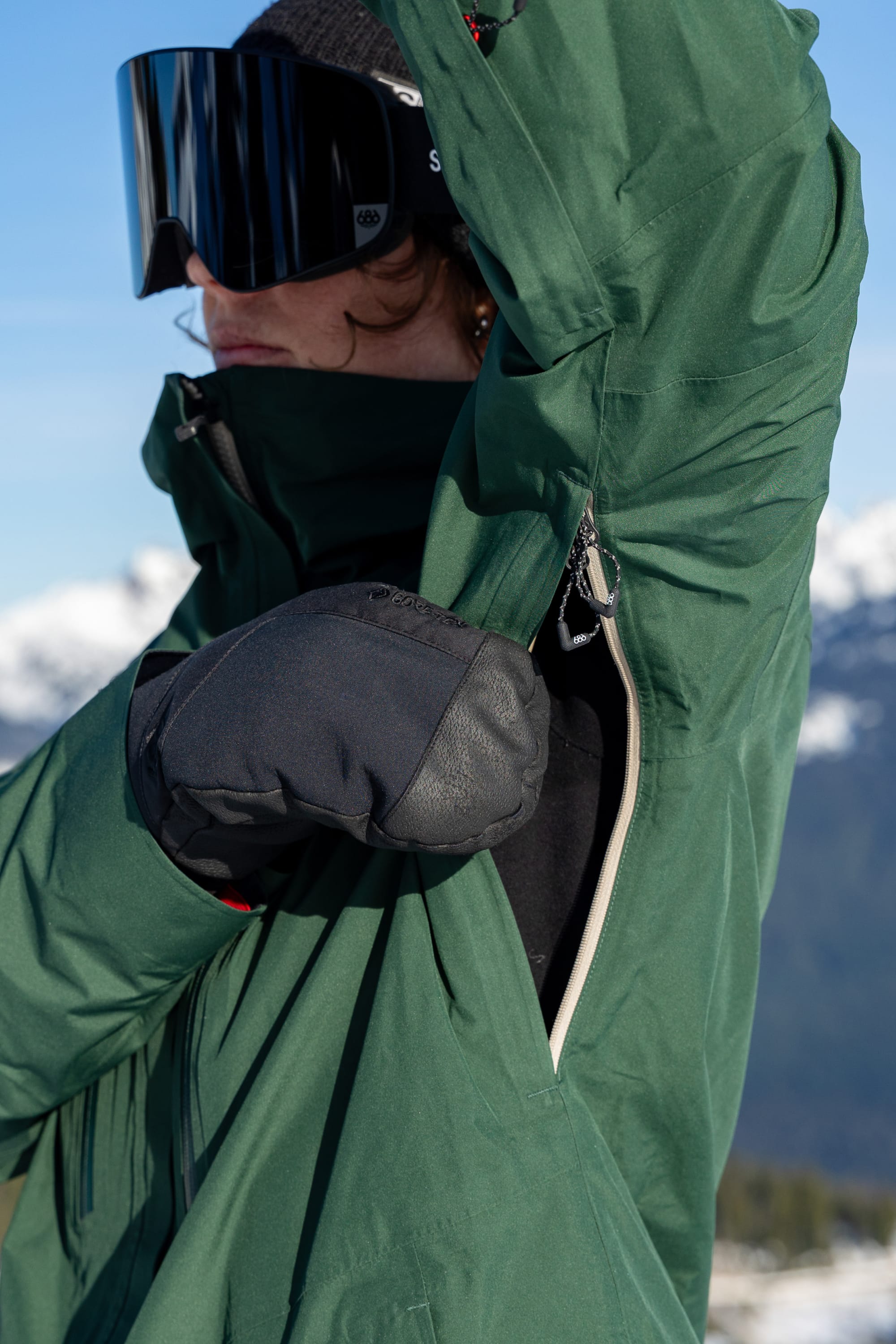 A person wearing the 686 Women’s GORE-TEX® Skyline Thermagraph® Jacket opens its underarm zipper for ventilation, with snowy mountains and a blue sky in the background.