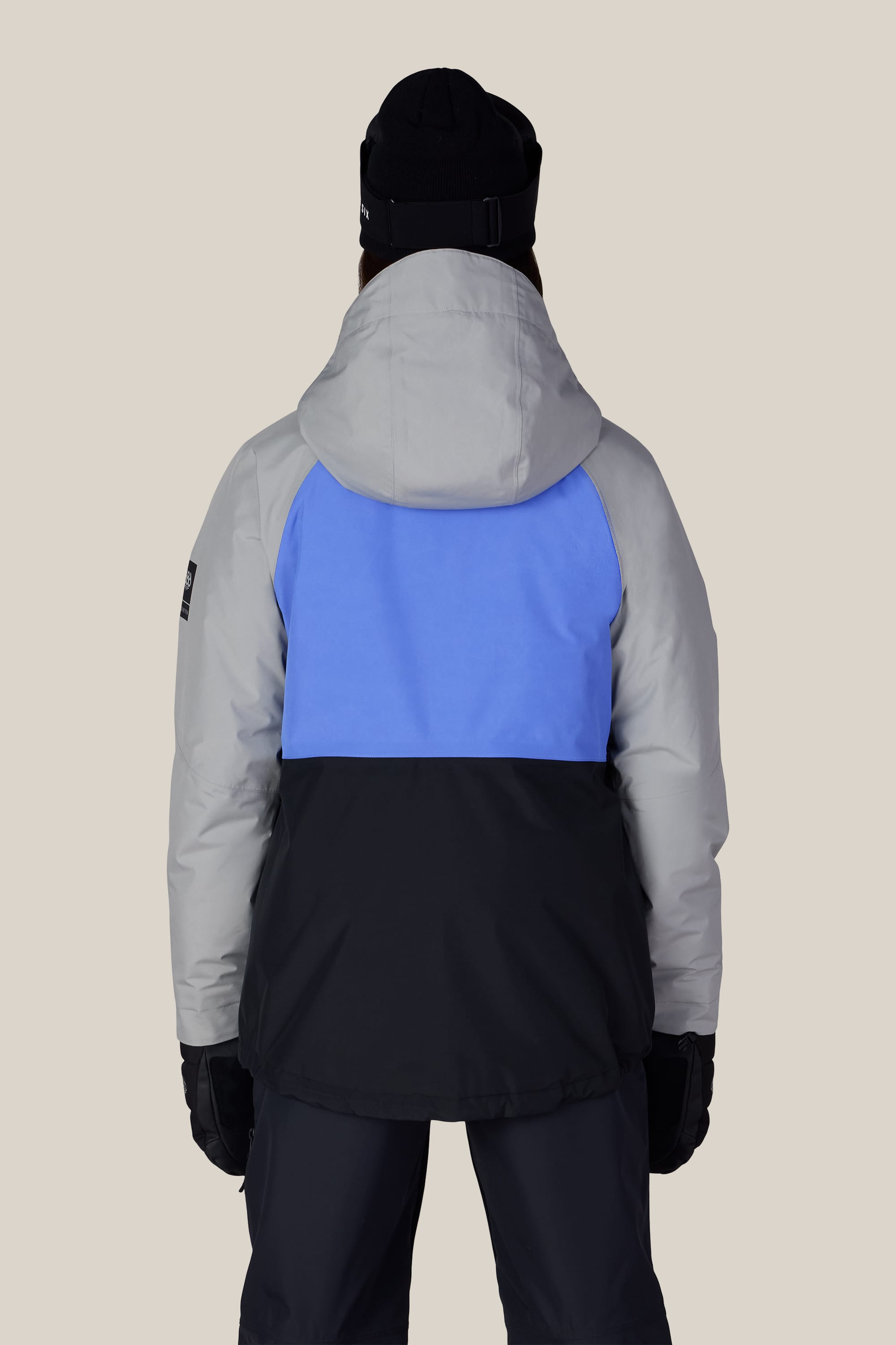 A person is seen from the back, standing against a plain background and wearing the 686 Women’s GORE-TEX® Willow™ Insulated Jacket from 686—a color-blocked style in gray, blue, and black—paired with dark pants, a black beanie, and gloves.