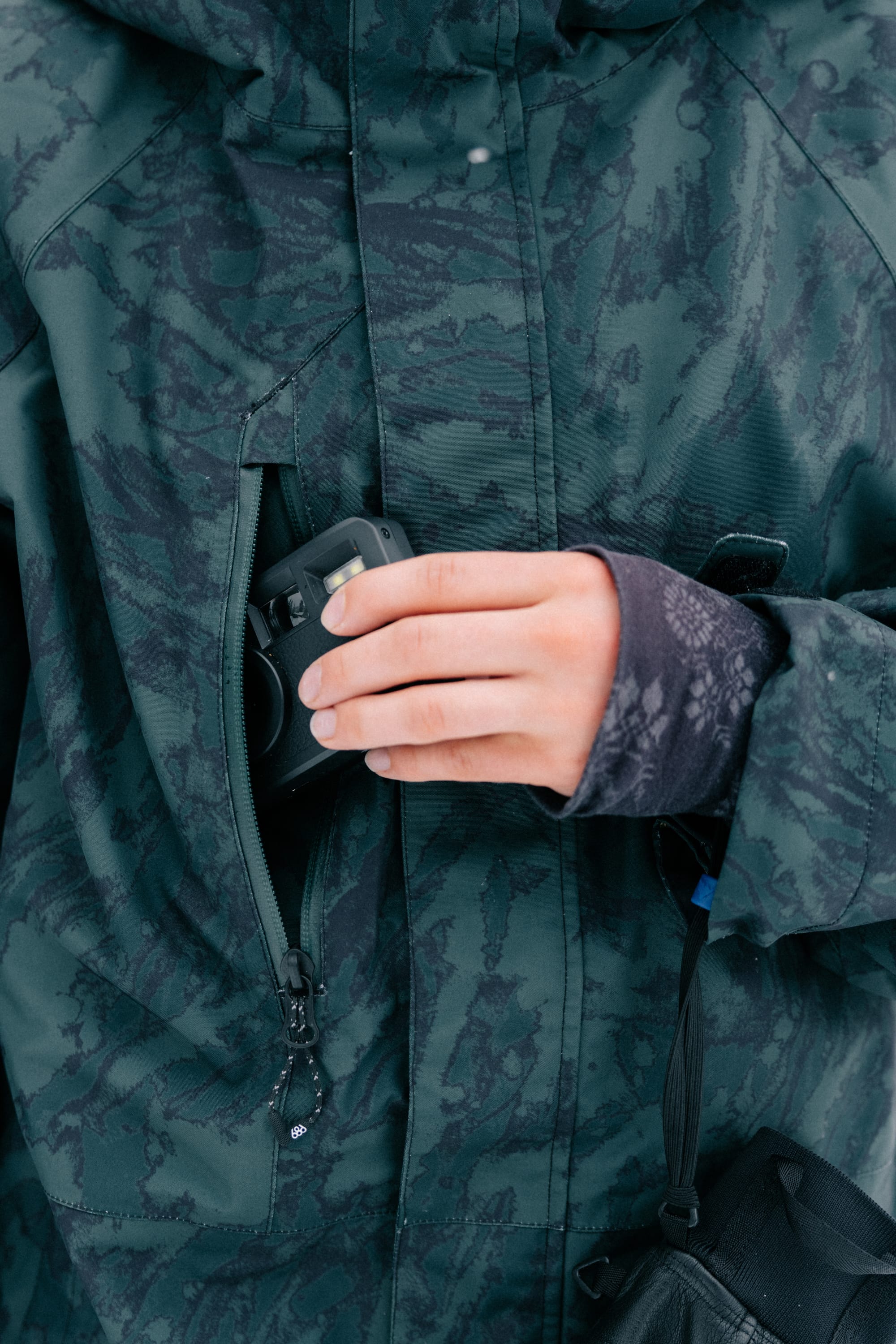 Wearing the 686 Women’s GORE-TEX® Willow™ Insulated Jacket, a person places a small black camera into the jacket’s zippered chest pocket, with only their hand and torso visible.
