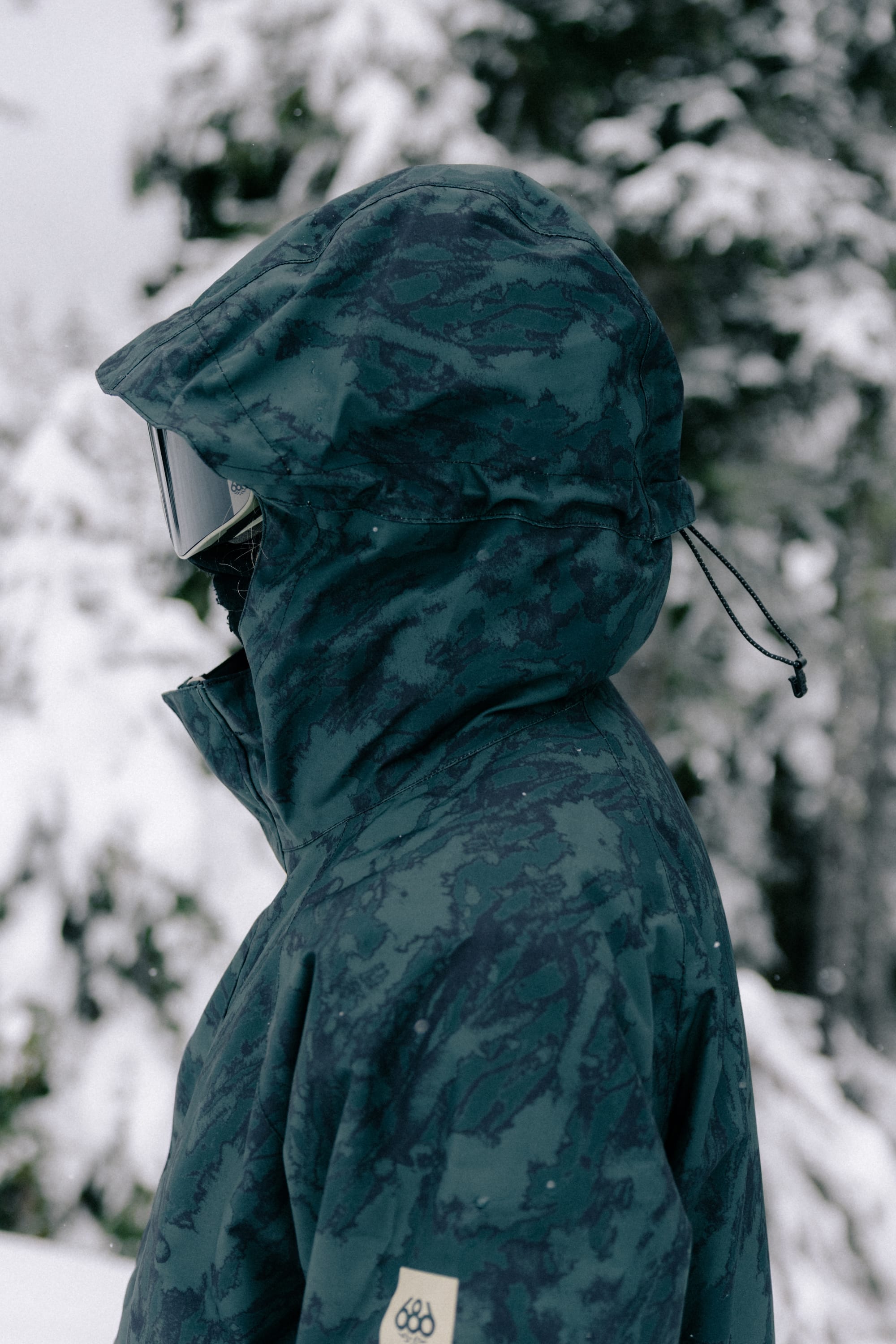Wearing the 686 Women’s GORE-TEX® Willow™ Insulated Jacket, a person stands outside in a snowy forest, facing left. The blue-green camouflage jacket from 686 is paired with ski goggles, while snow-covered trees blur in the background.