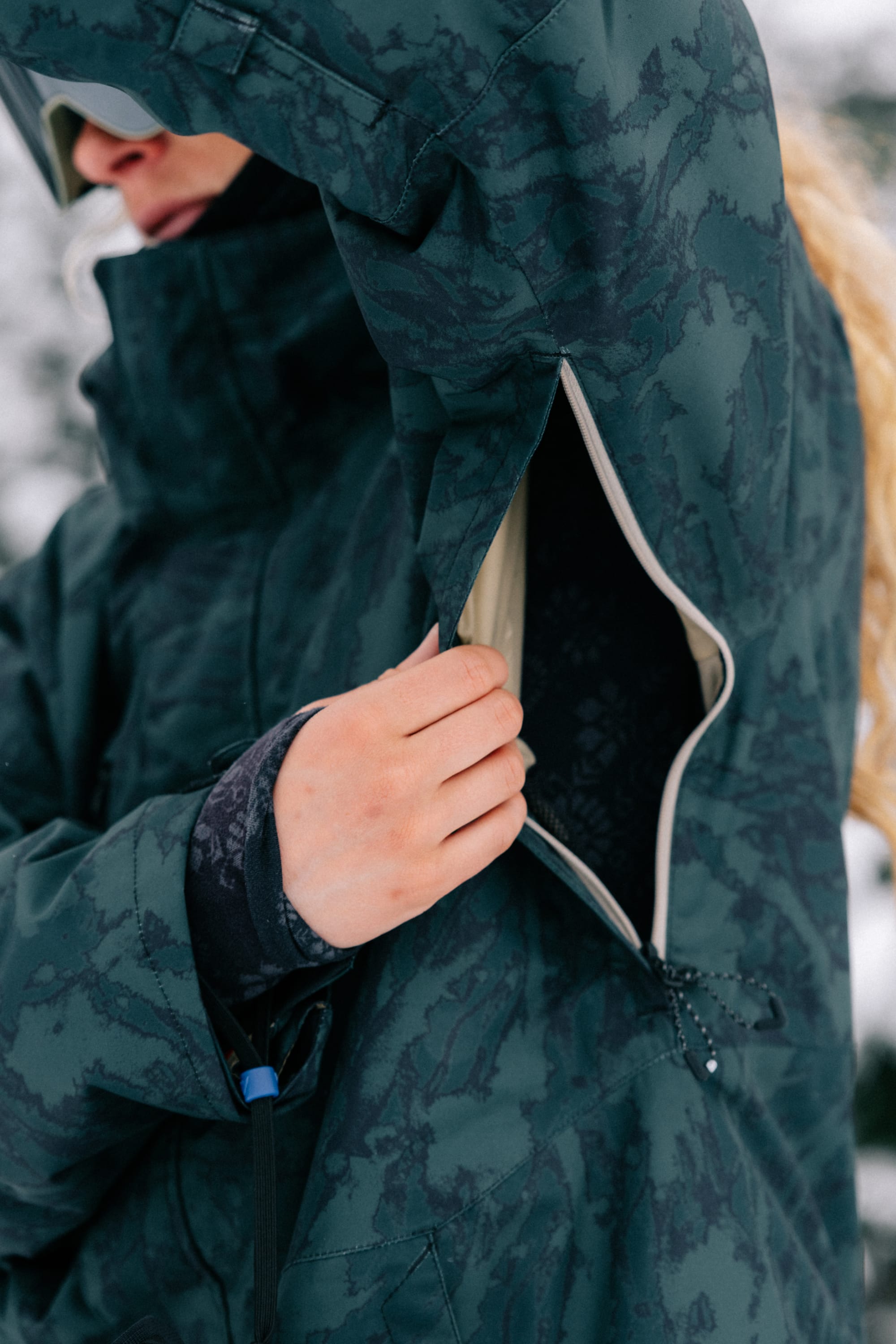 Wearing the 686 Women’s GORE-TEX® Willow™ Insulated Jacket in a patterned green design, a person opens the armpit zipper for ventilation, their hand partially inside the jacket while snow is visible in the blurred background.