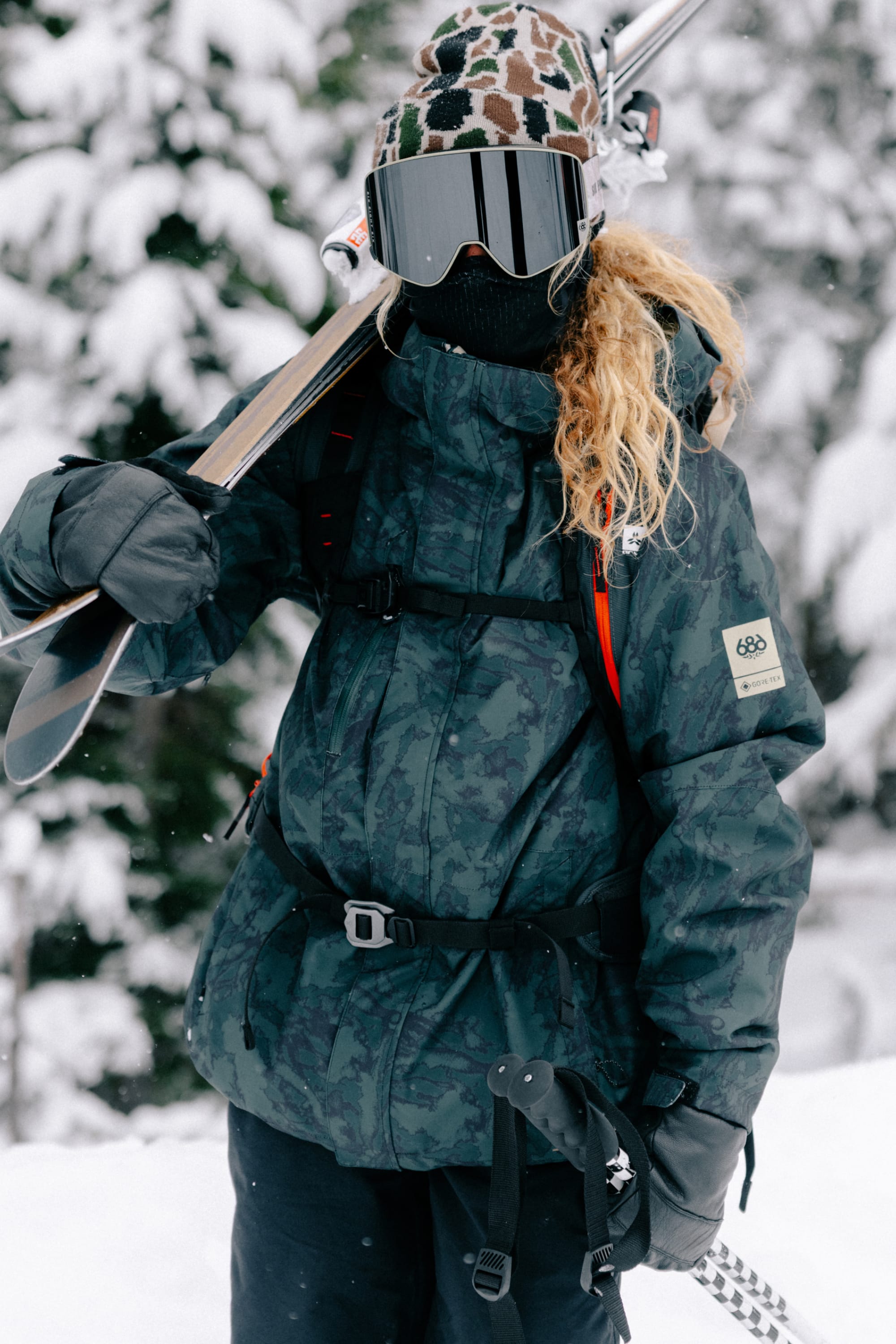 A person in winter gear stands outdoors in the snow, holding skis and ski poles. They wear a patterned beanie, dark ski goggles, gloves, and the 686 Women’s GORE-TEX® Willow™ Insulated Jacket from 686. Snow-covered trees can be seen in the background.