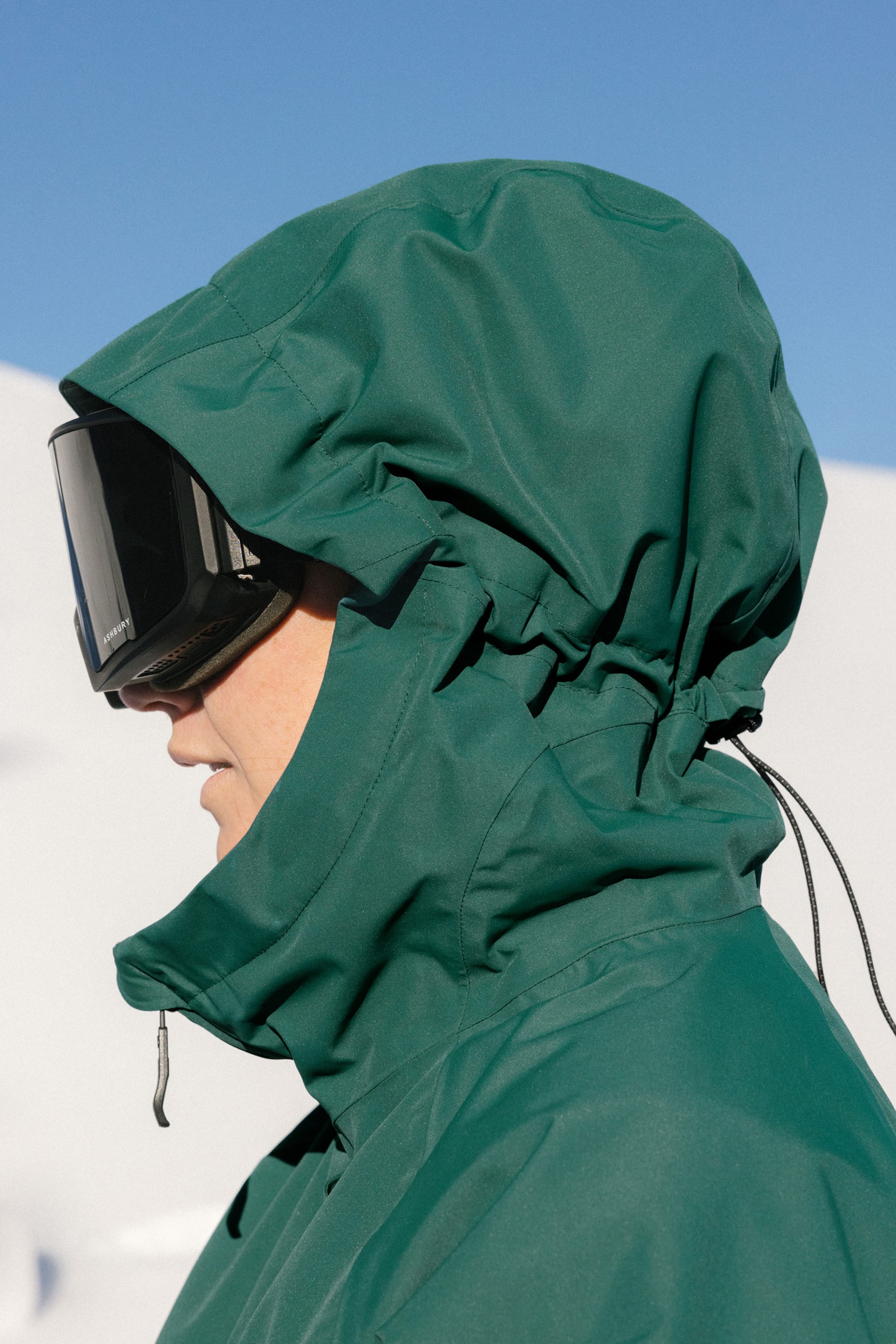 A person wears the 686 Womens Whisper Insulated Jacket in green and black ski goggles, standing outside in a snowy landscape under a clear blue sky.