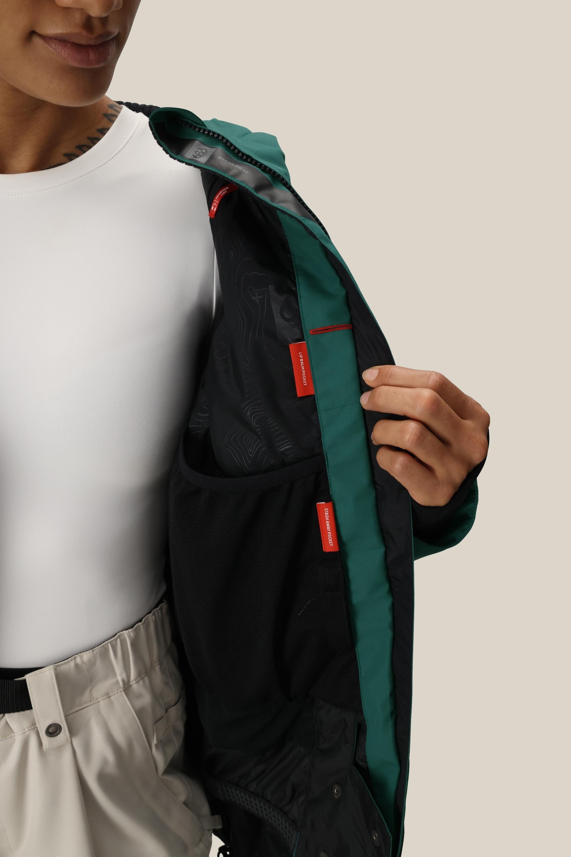A person wearing a white shirt and beige pants opens the 686 Womens Whisper Insulated Jacket in dark green, revealing its black lining with multiple red tags and mesh pockets.
