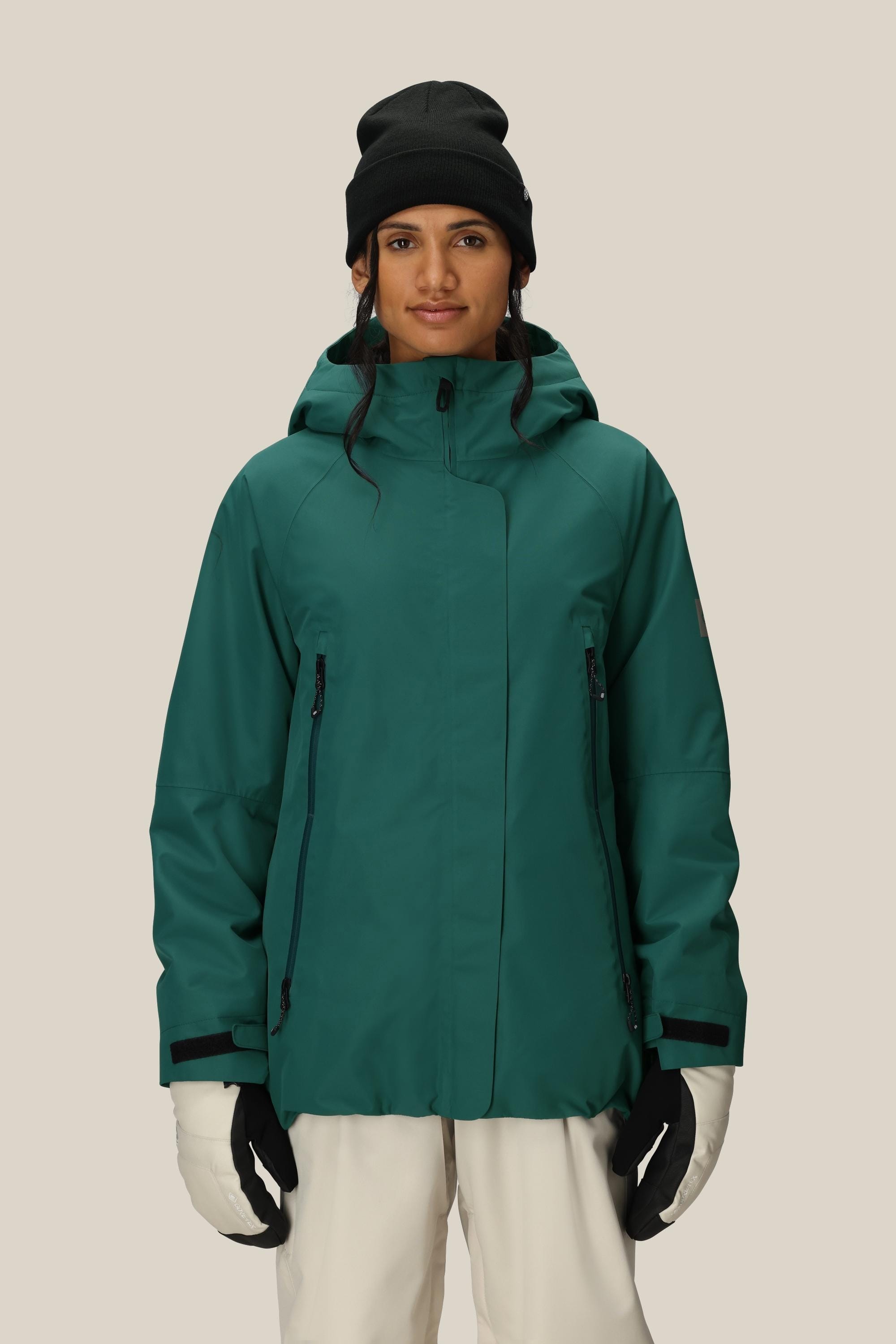 A woman wears the 686 Womens Whisper Insulated Jacket in dark green with a hood, paired with white gloves, white pants, and a black beanie, standing against a plain beige background.