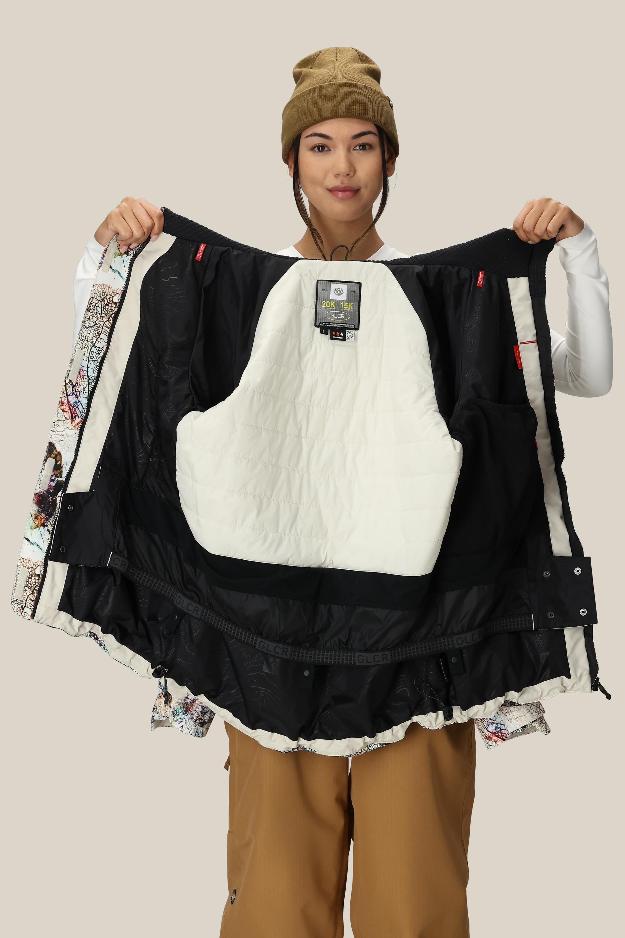 A person in a brown beanie and pants holds open the 686 Womens Whisper Insulated Jacket by 686, revealing its quilted white and black interior, against a plain light background.