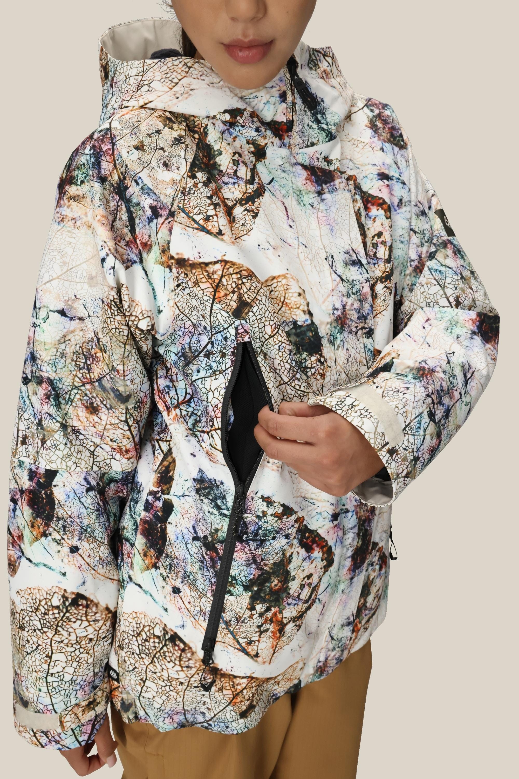 A person wearing the 686 Womens Whisper Insulated Jacket featuring a multicolored leaf pattern opens its zippered chest pocket. The hooded, waterproof jacket by 686 is shown from shoulders to mid-thigh against a plain background.