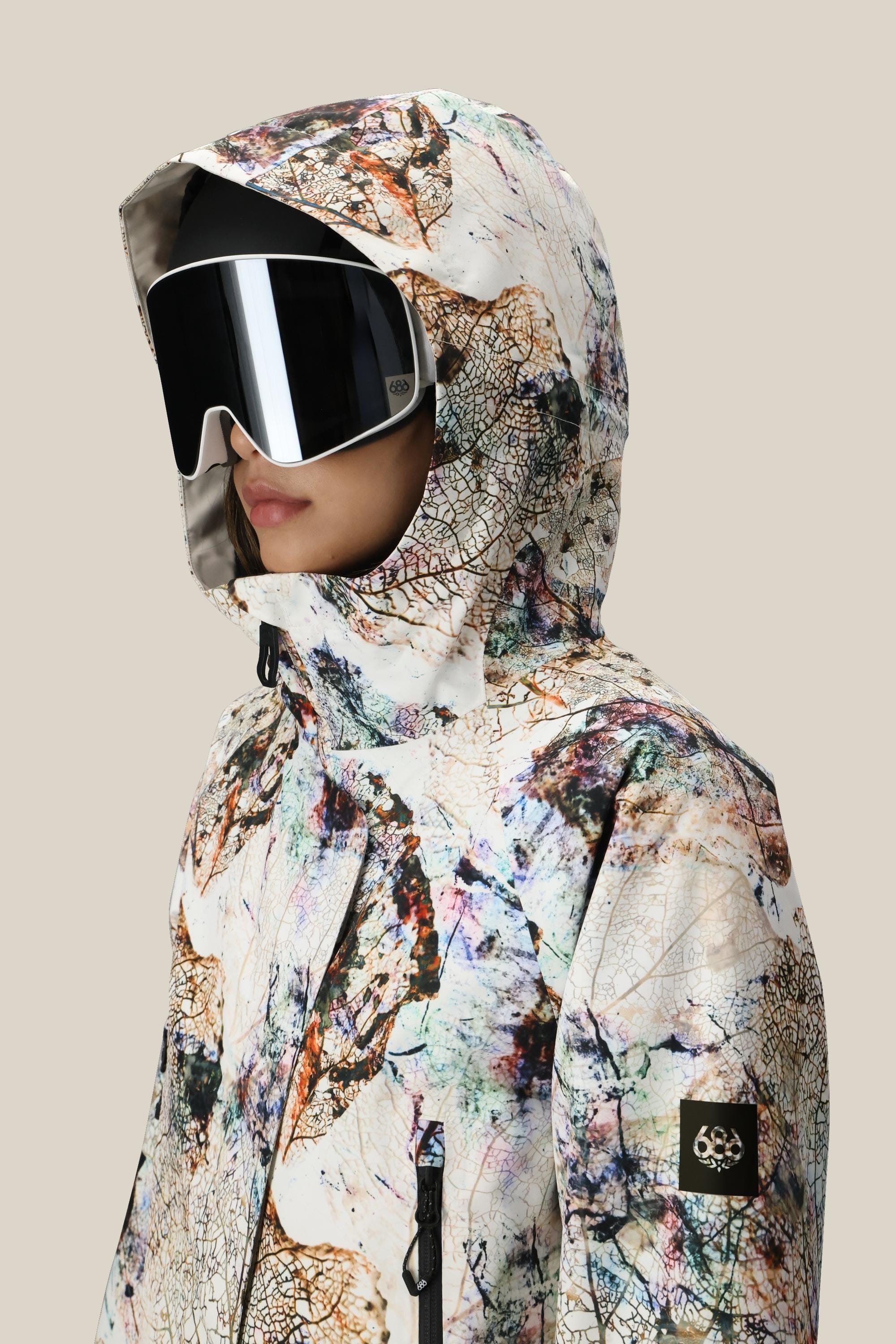 A person wearing the 686 Womens Whisper Insulated Jacket with a nature-inspired abstract print and large black ski goggles, facing left. The hooded polyester jacket features a sleeve logo patch, shown against a neutral background.