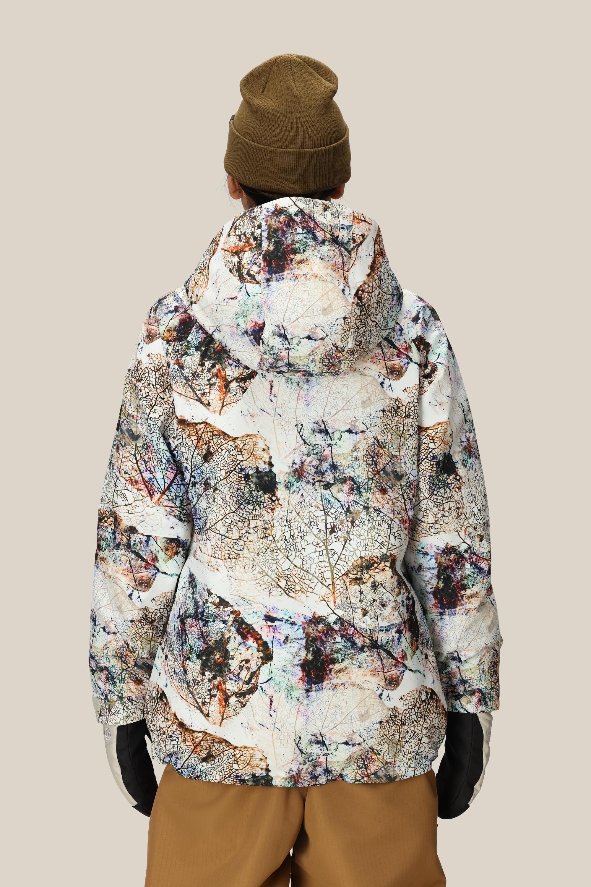 A person stands with their back to the camera, wearing the 686 Womens Whisper Insulated Jacket by 686 featuring an abstract nature pattern, paired with a brown beanie, brown pants, and black gloves against a plain beige background.