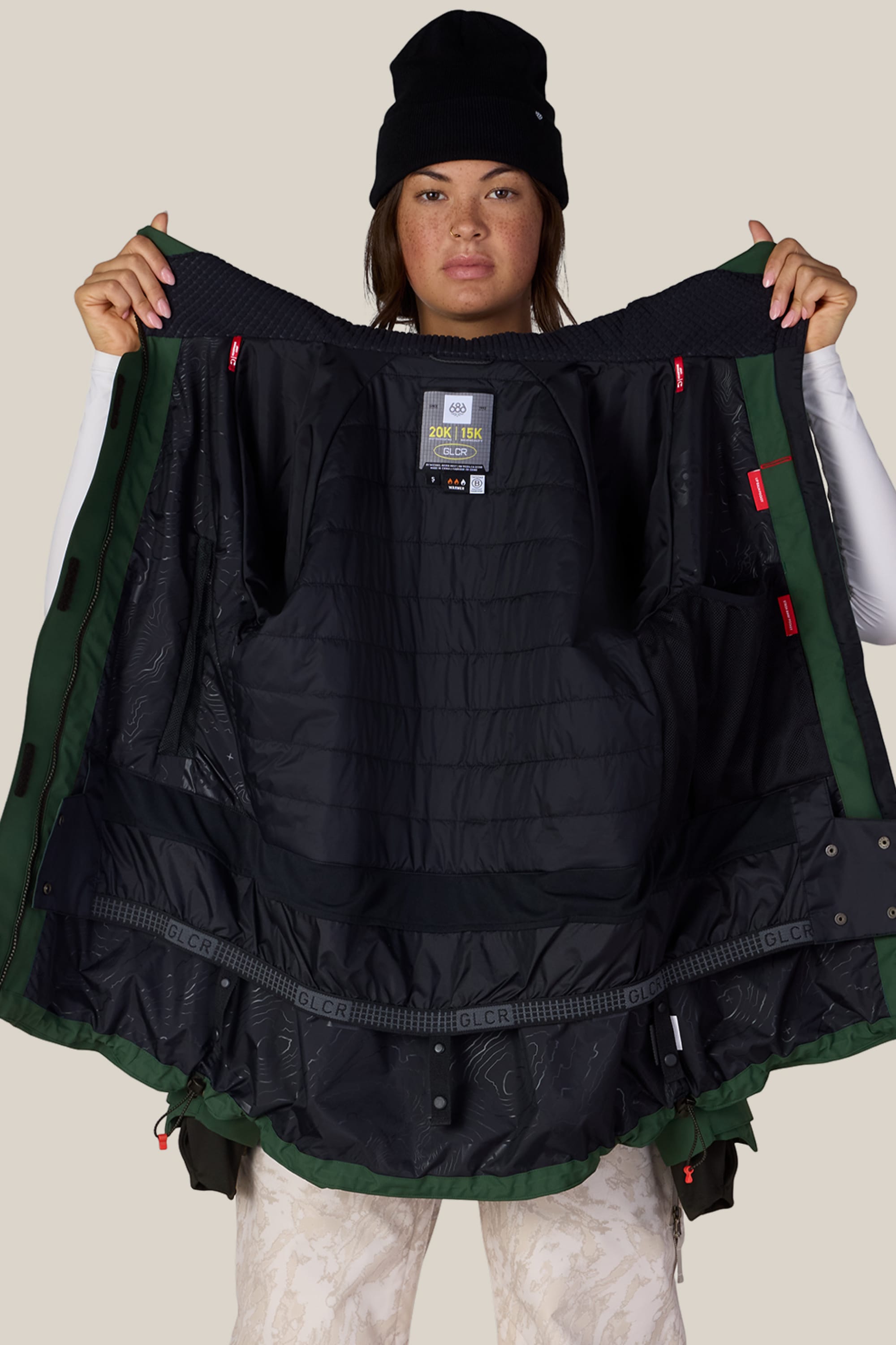 A person wearing a black beanie and patterned pants holds open the 686 Women’s Whisper™ Insulated Jacket by 686, displaying its quilted interior and multiple pockets.