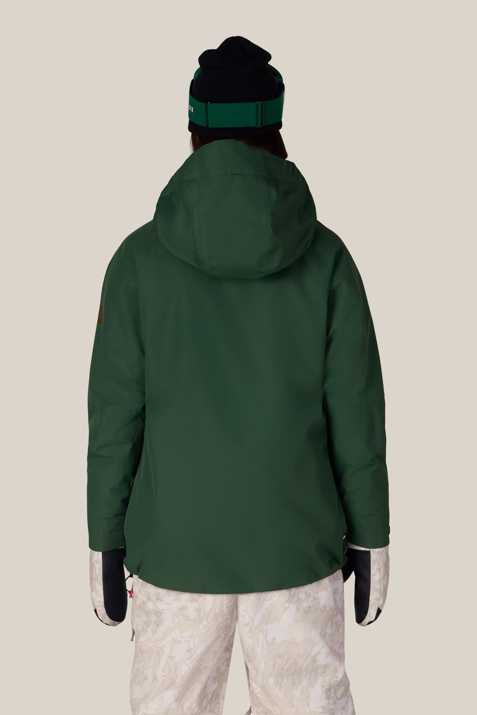 Wearing the 686 Women’s Whisper™ Insulated Jacket by 686 in dark green, along with a black and green beanie, white patterned snow pants, and black and white mittens, this person is standing with their back to the camera against a plain beige background.