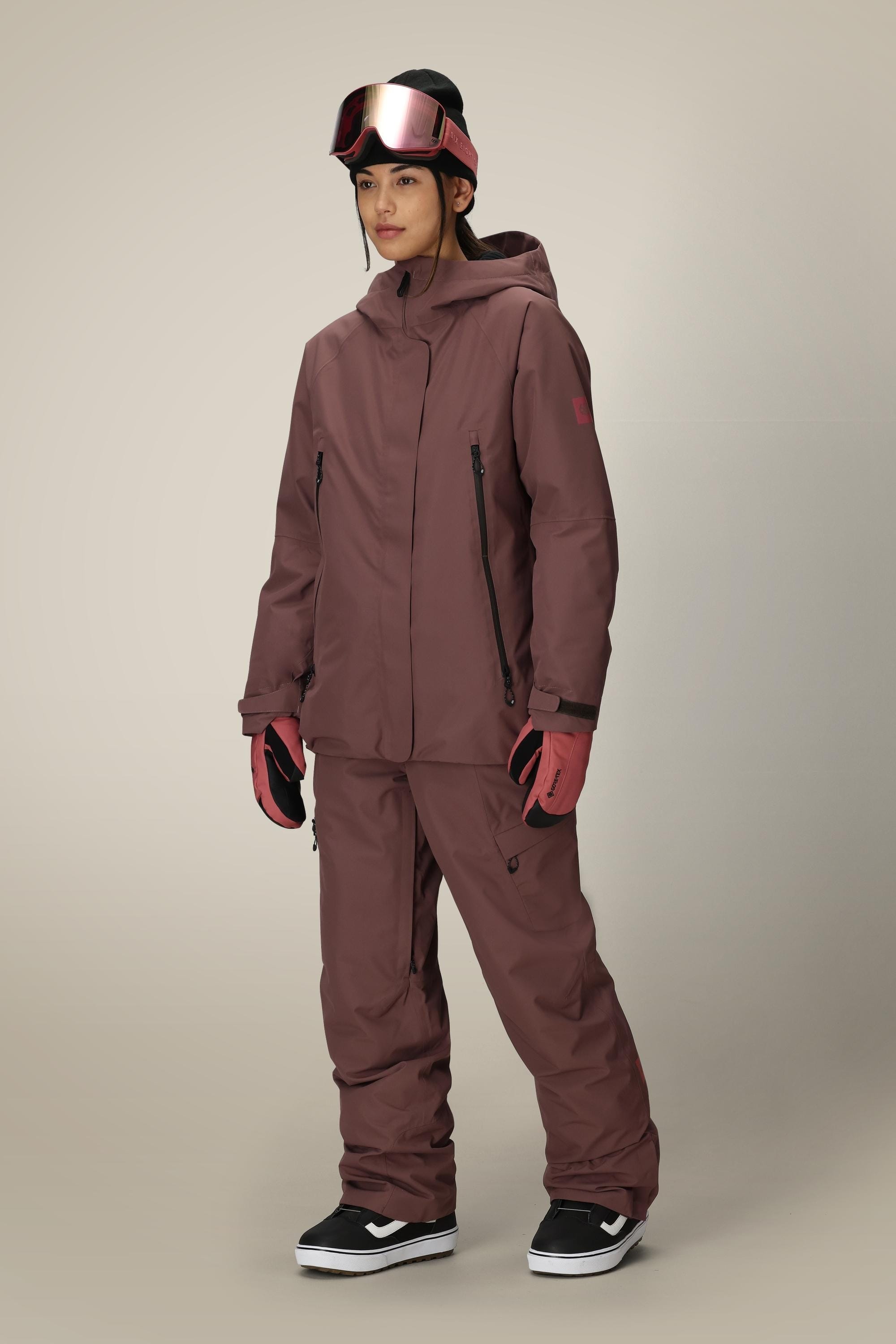 A person wearing a maroon 686 Womens Whisper Insulated Jacket and pants, pink gloves, black snow boots, and pink ski goggles stands against a plain light background.