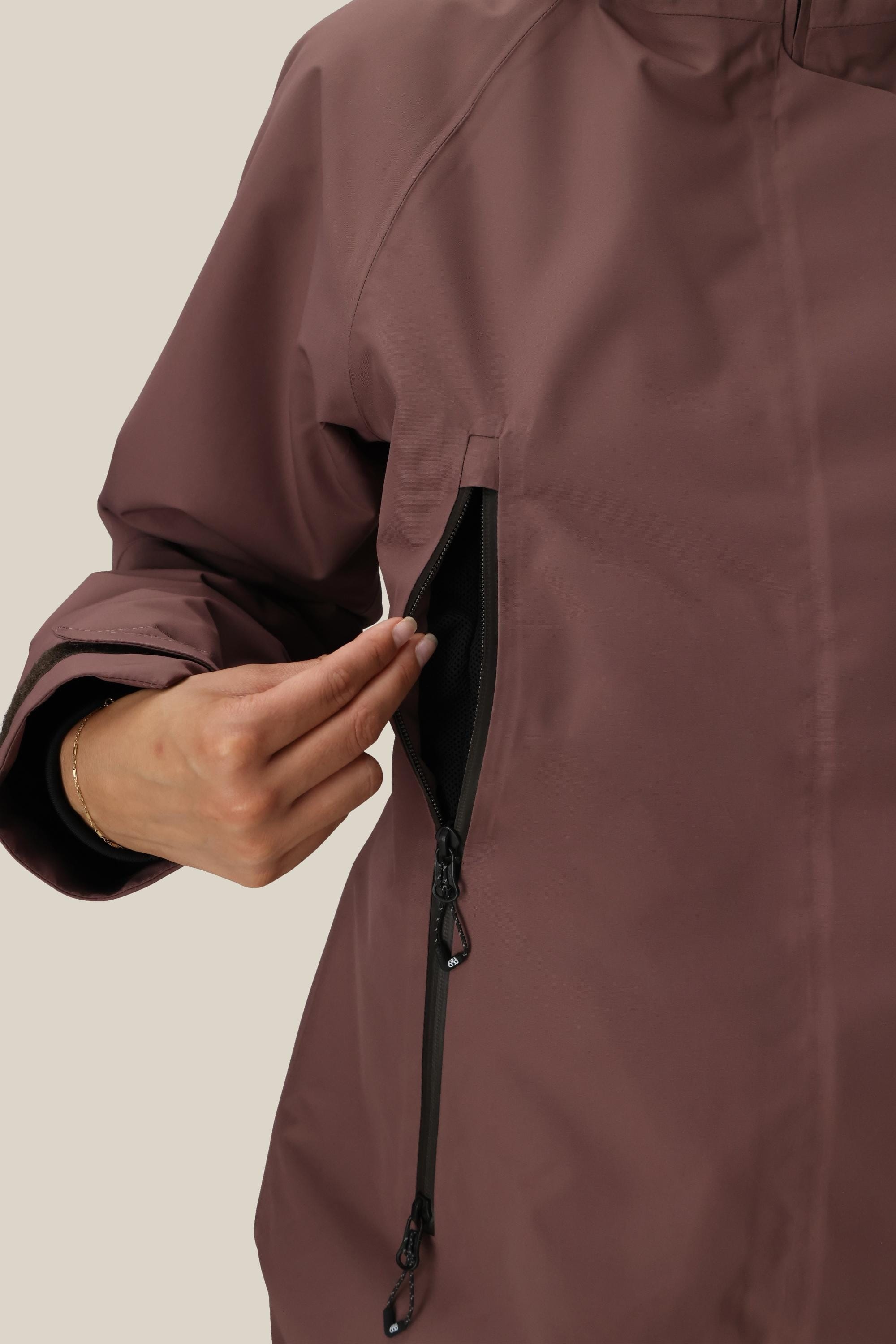 A person wearing the 686 Womens Whisper Insulated Jacket in mauve uses their right hand to unzip a vertical pocket on the left side, revealing the inner mesh lining.