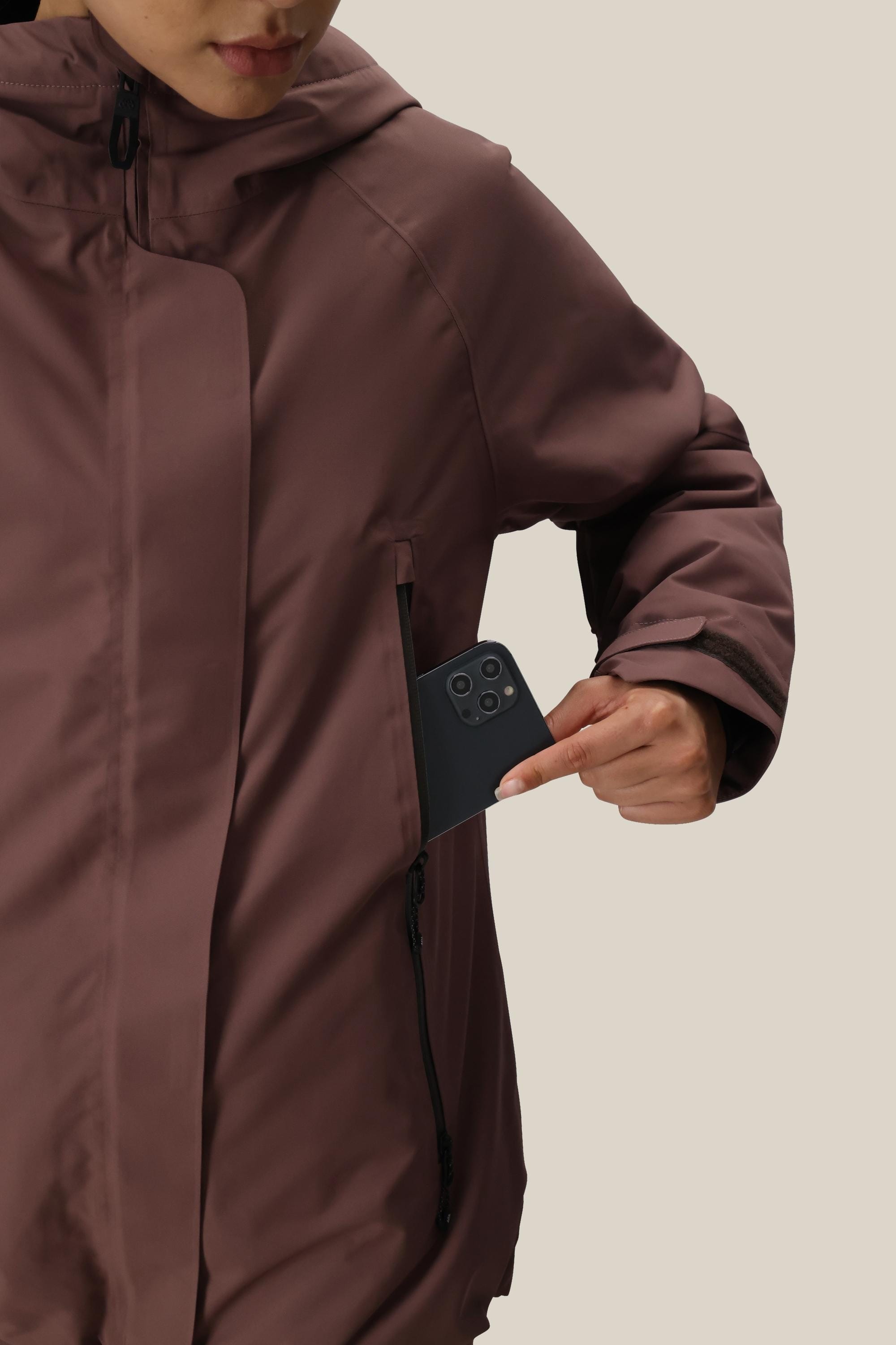 A woman wearing the 686 Women’s Whisper Insulated Jacket in brown slides a black smartphone into its zippered chest pocket. The background is plain and light-colored.